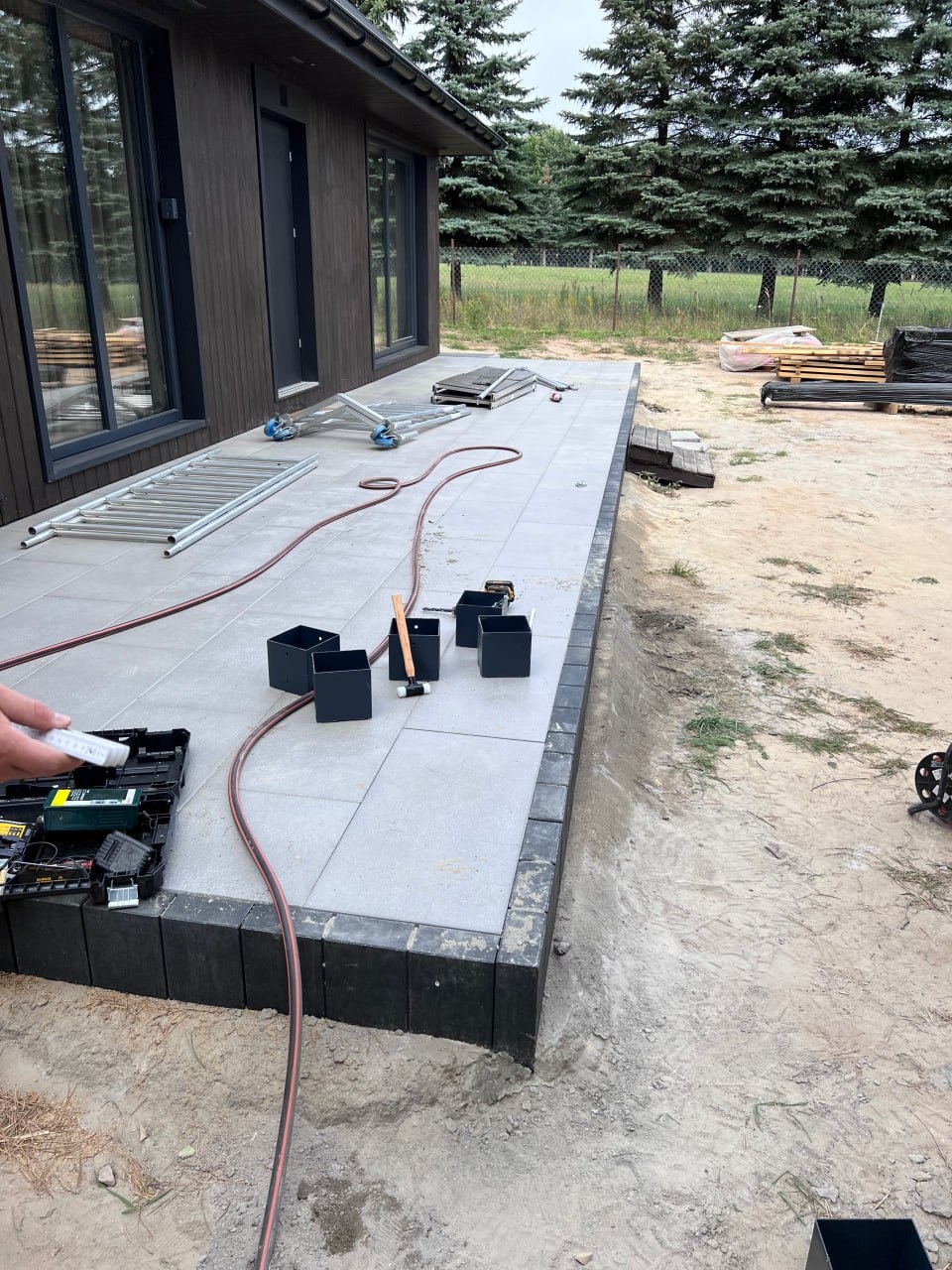 Montaż tarasu z szarych płyt i czarnych obrzeży przy brązowym domu. Widoczne narzędzia i akcesoria montażowe. Prace w toku, teren wokół w piasku.