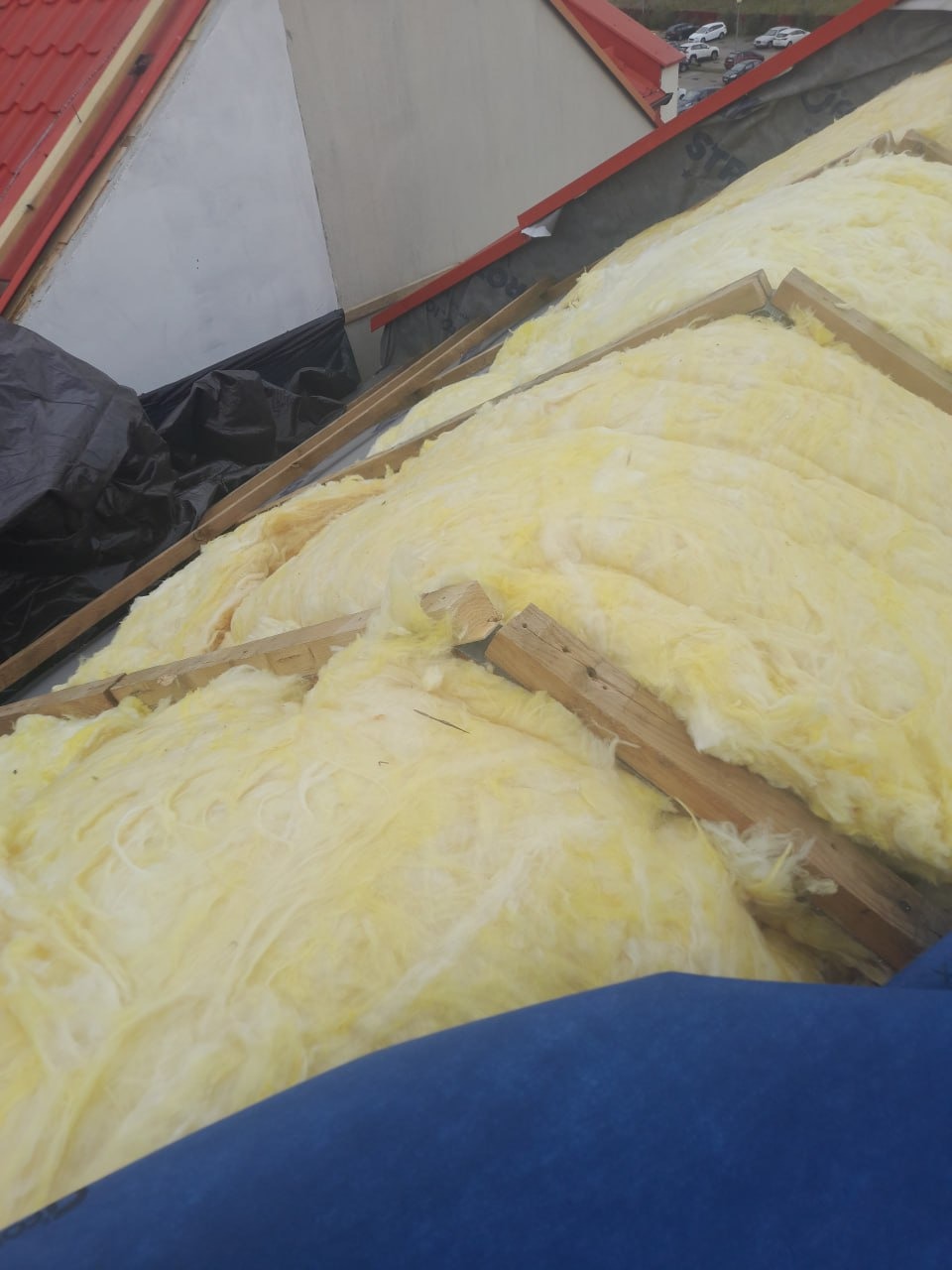 Żółta wełna mineralna między krokwiami dachu, widok z góry. Częściowo widoczna membrana dachowa i czerwona dachówka. Ocieplenie poddasza w trakcie realizacji.