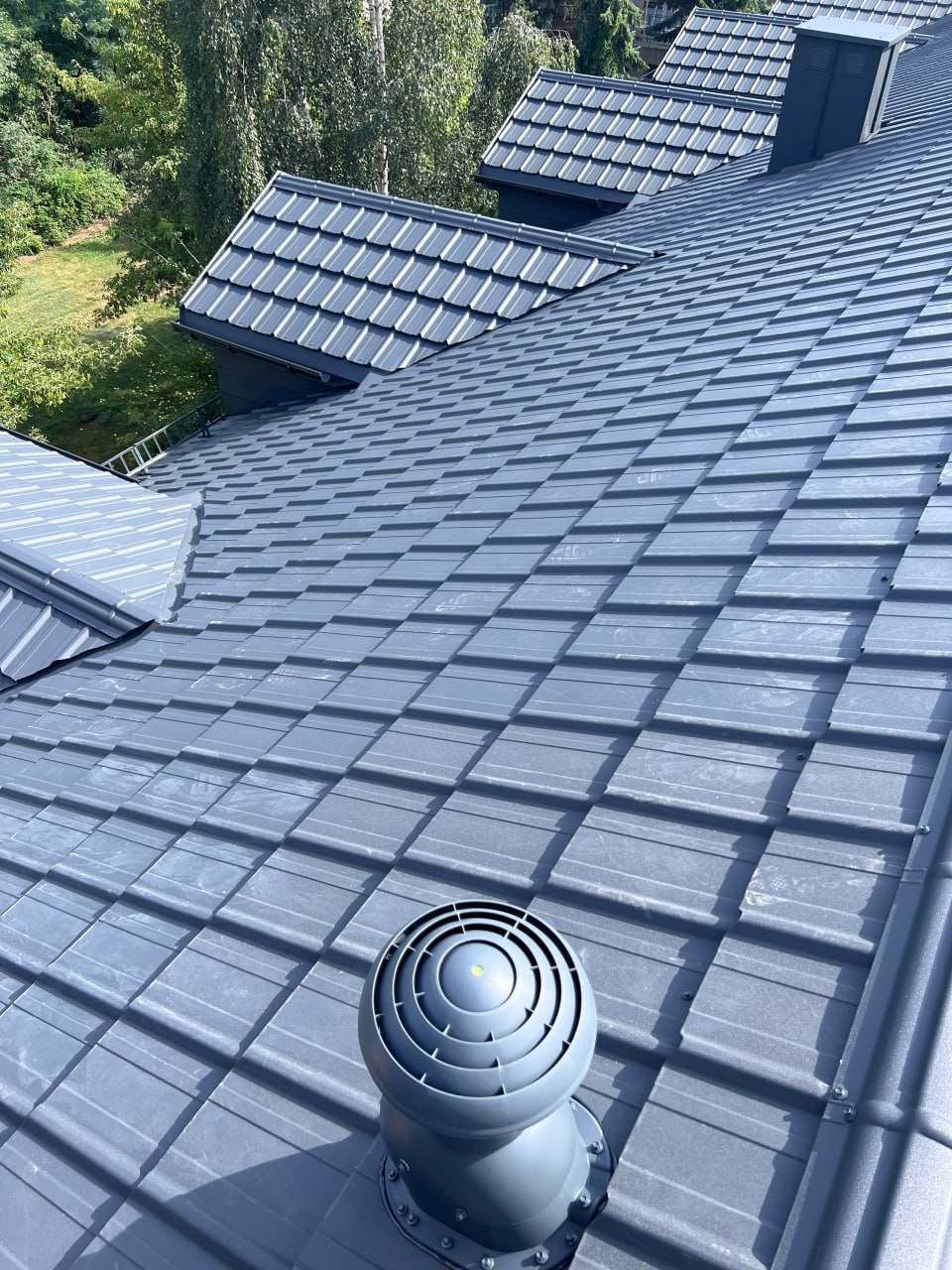 Szary dach z blachy trapezowej z wentylatorem wywiewnym. Widok z góry, w tle zieleń drzew. Nowoczesne pokrycie dachowe.