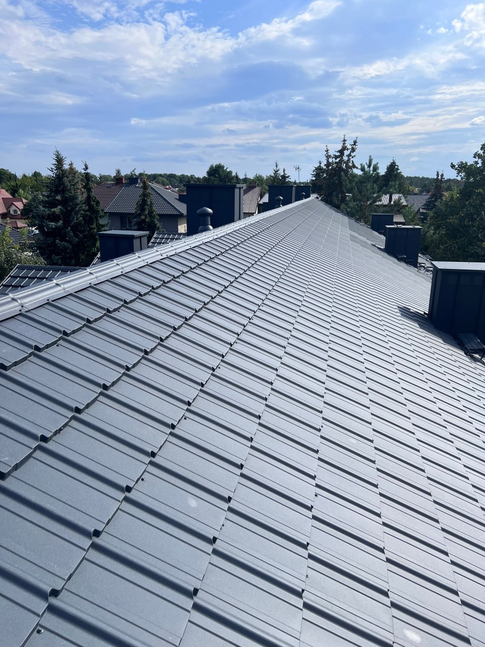 Szary dach z blachy trapezowej po kryciu w Warszawie, widok z góry na tle domów i drzew. Nowe pokrycie dachu, regularny wzór blachy.