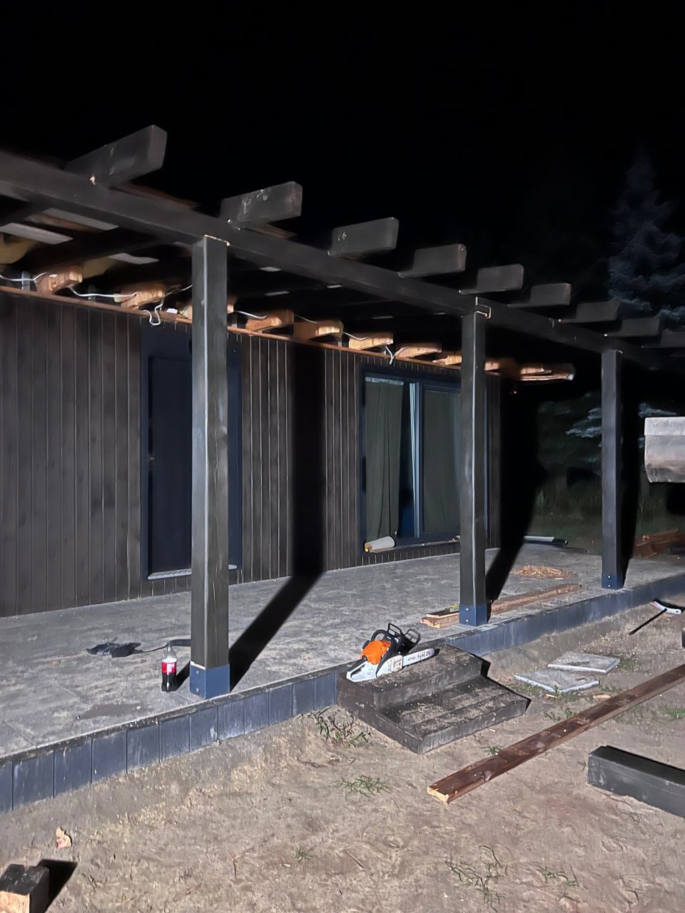 Ciemna pergola w trakcie budowy, z widocznymi elementami konstrukcyjnymi i piłą łańcuchową, nocne ujęcie z bliska, oświetlenie punktowe, częściowo ukończona konstrukcja.