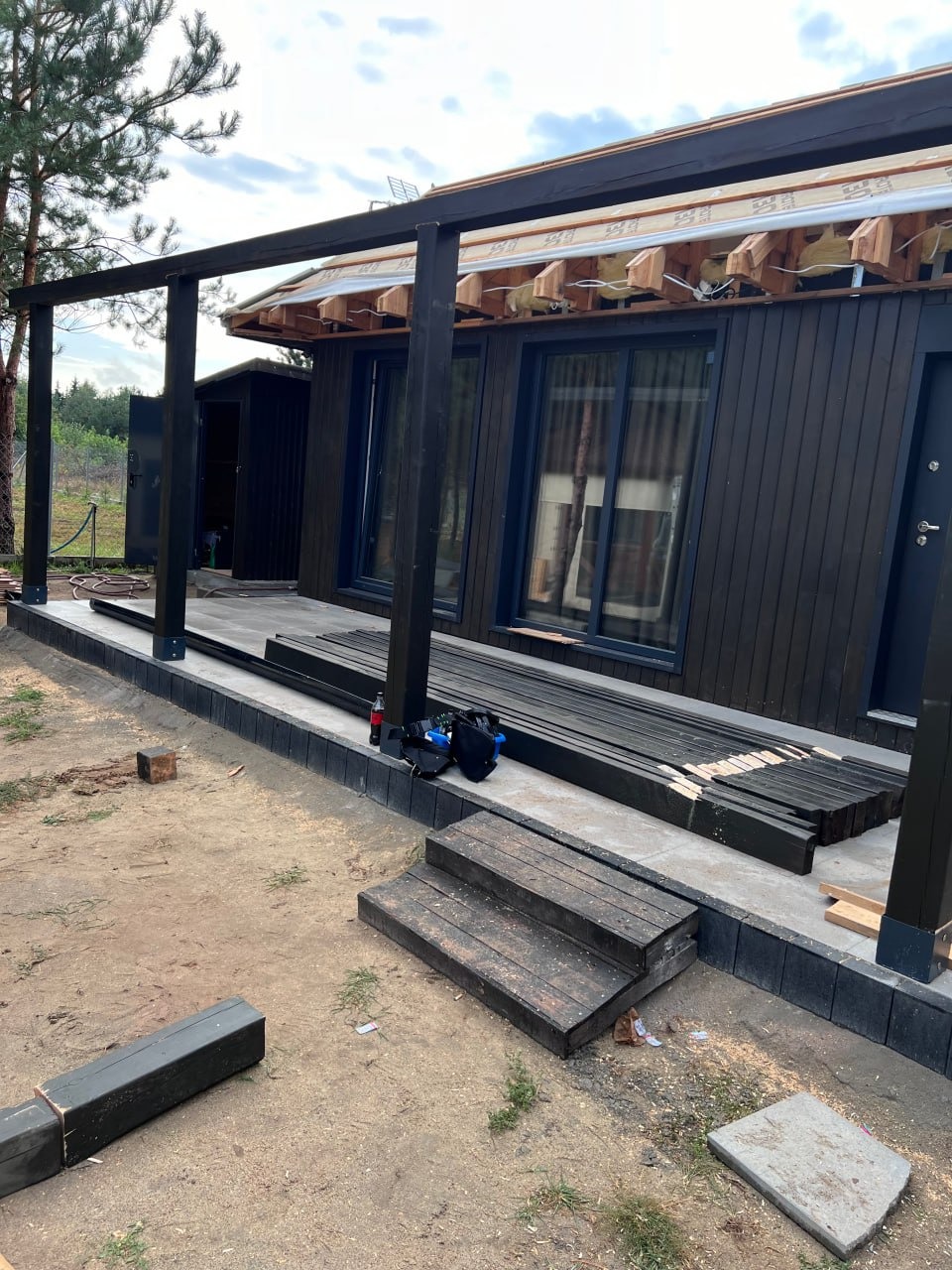 Dom z ciemną elewacją w trakcie budowy z widoczną konstrukcją dachu i elementami drewnianymi przygotowanymi do montażu na zewnątrz budynku.