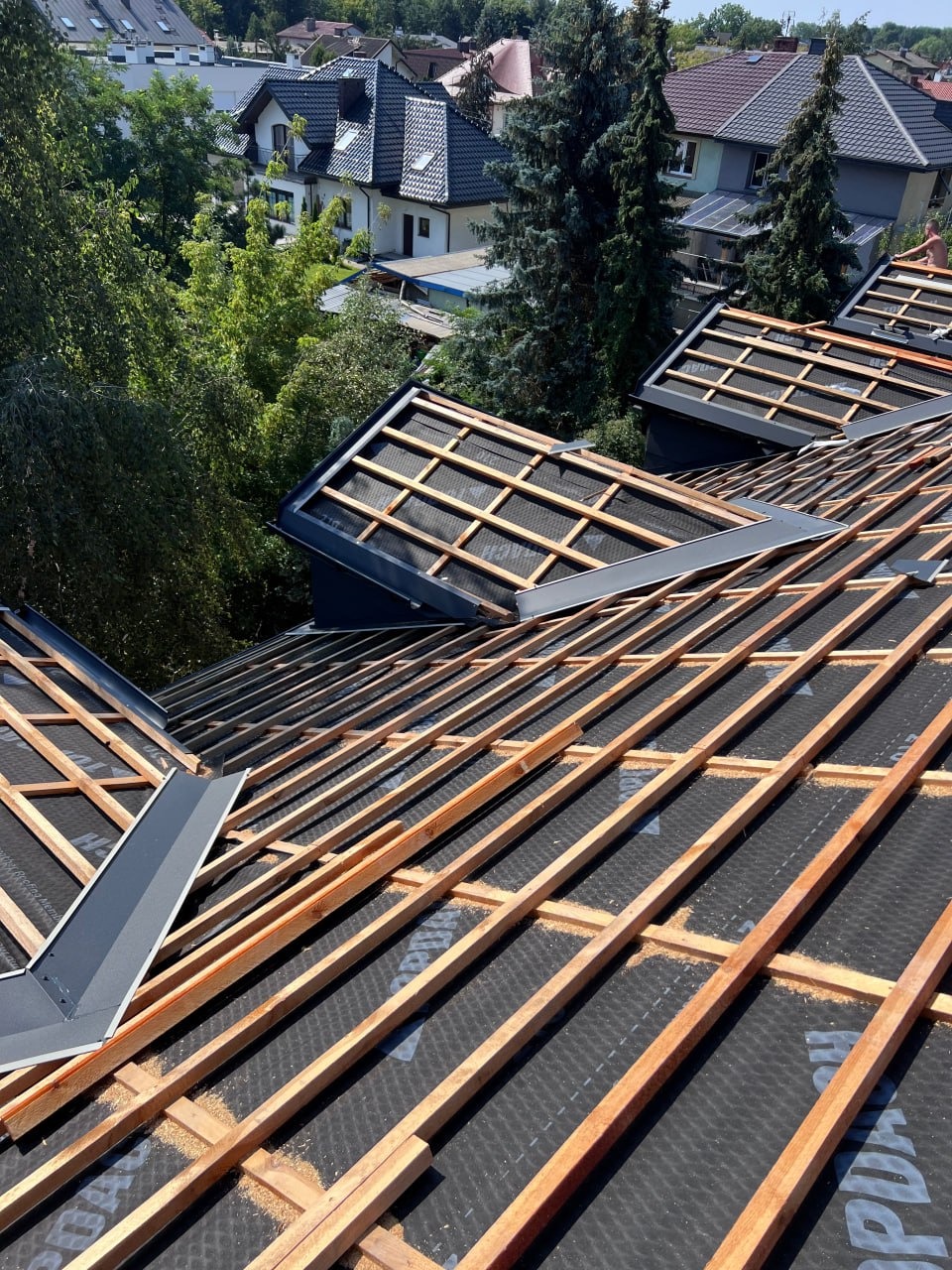 Konstrukcja dachu z łatami i membraną, widok z góry na tle zabudowy jednorodzinnej i zieleni, oświetlona słońcem.  Szczegółowo widoczne obróbki blacharskie.