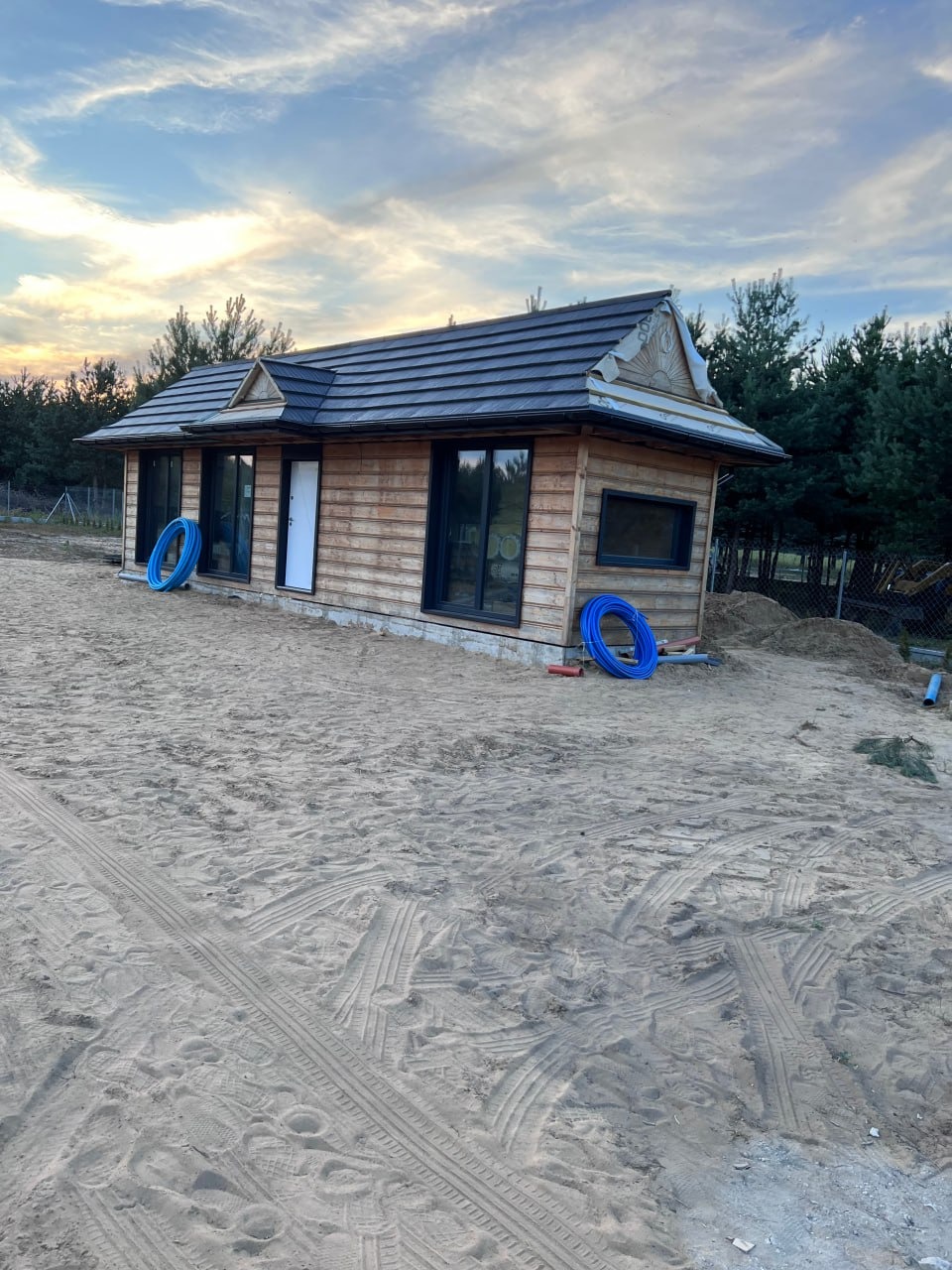 Drewniany domek z ciemną dachówką na piaszczystym terenie. Widoczne niebieskie rury i las w tle. Dom z białymi drzwiami i czarnymi ramami okiennymi.