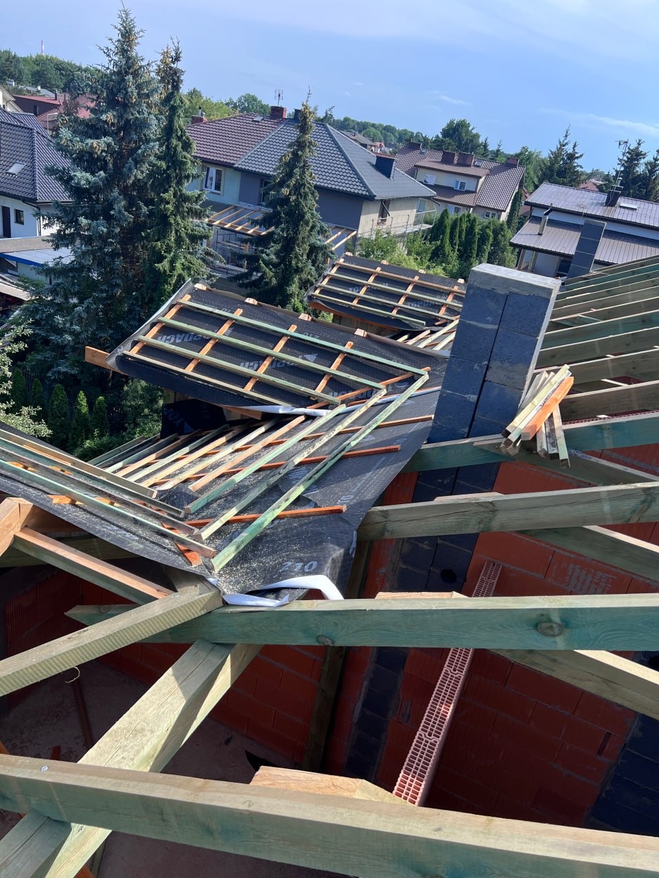 Konstrukcja dachu w trakcie budowy z widocznymi elementami drewnianymi, membraną dachową i kominem w tle zabudowy jednorodzinnej.