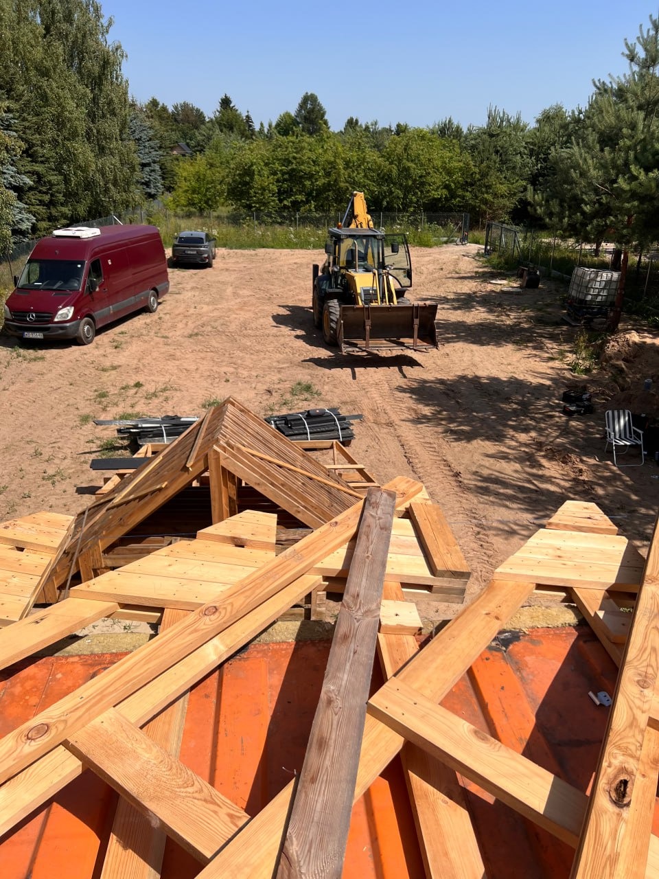 Drewniana konstrukcja dachu w trakcie budowy, widok z góry. W tle czerwony van, koparka i drzewa. Prace dekarskie w toku, elementy konstrukcyjne na pomarańczowej membranie.