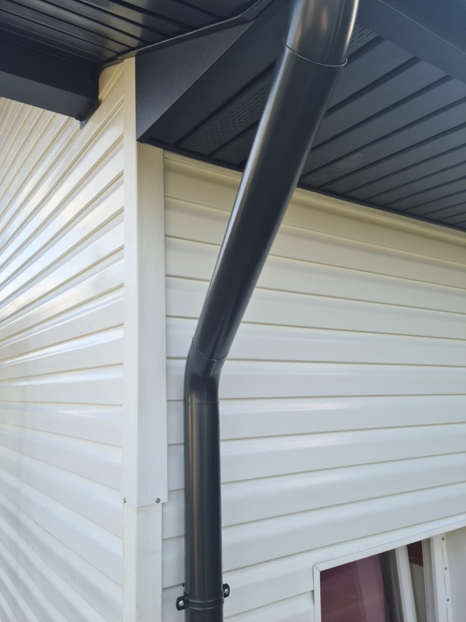 Szara rynna spustowa zamontowana na elewacji z białego sidingu, widok z dołu. Eleganckie wykończenie dachu i narożnika budynku.