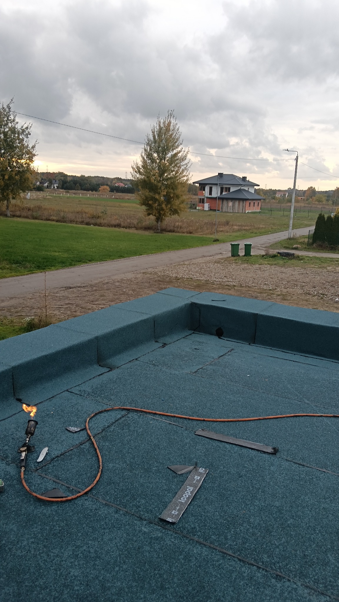 Prace dekarskie na płaskim dachu: palnik dekarski w użyciu, widoczne narzędzia i fragmenty papy. W tle krajobraz z budynkiem. Dach pokryty zieloną papą.