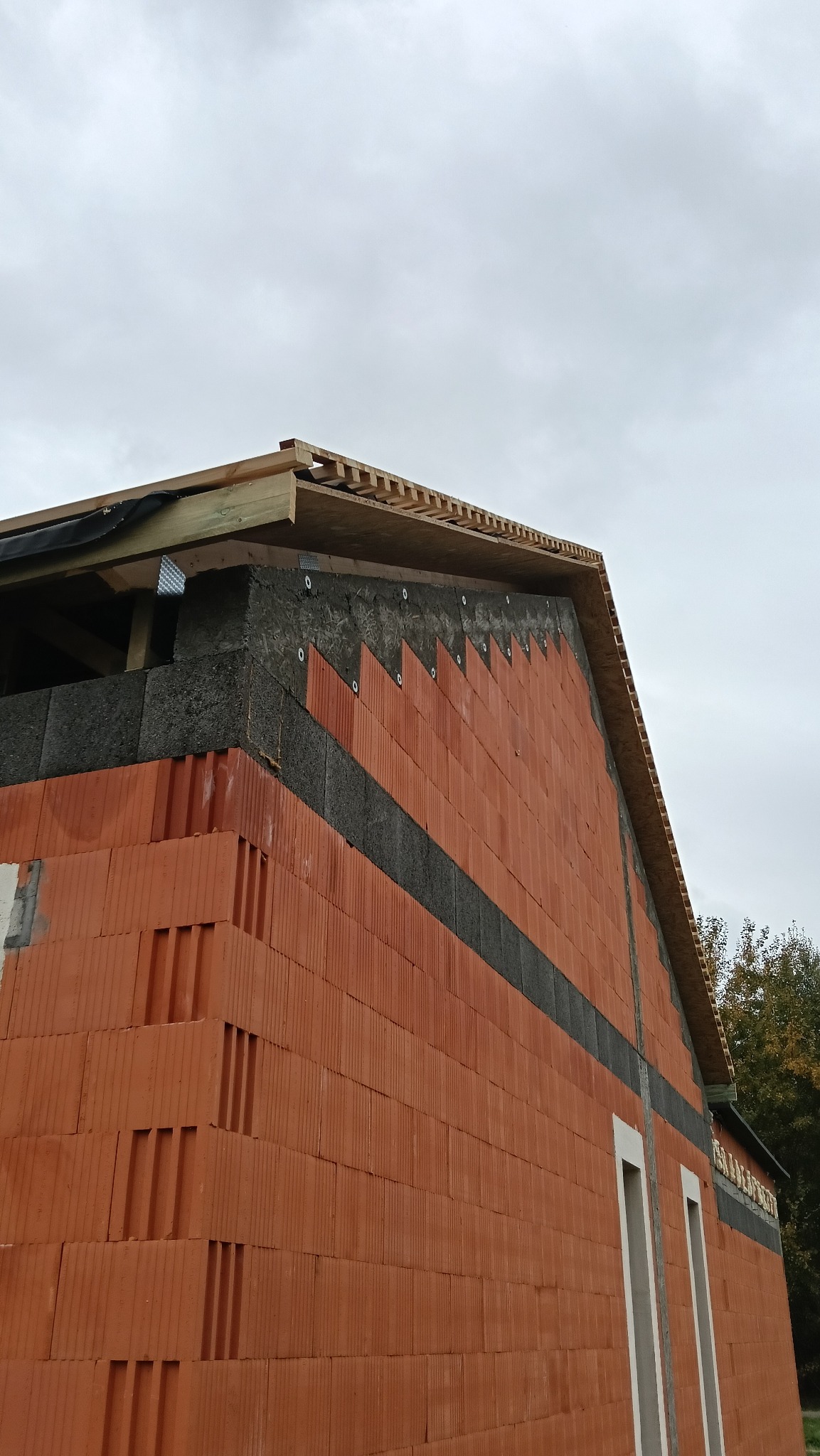 Budowa domu z czerwonej cegły, widoczne ocieplenie i fragment konstrukcji dachu. Ujęcie z dołu na fragment ściany szczytowej i okapu.