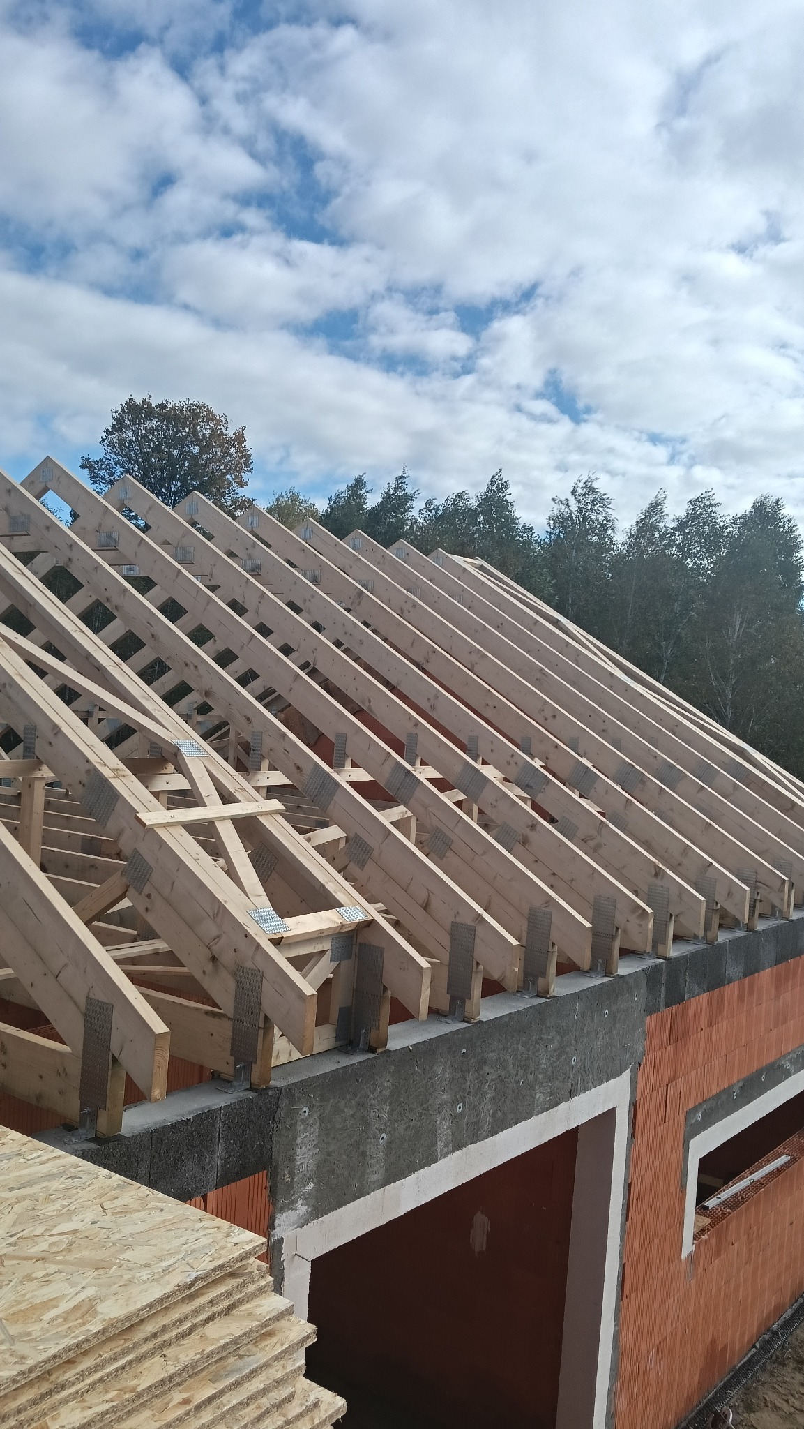 Konstrukcja więźby dachowej z drewna, zabezpieczona metalowymi łącznikami, na tle błękitnego nieba z chmurami. Widoczne elementy muru budynku z czerwonej cegły.