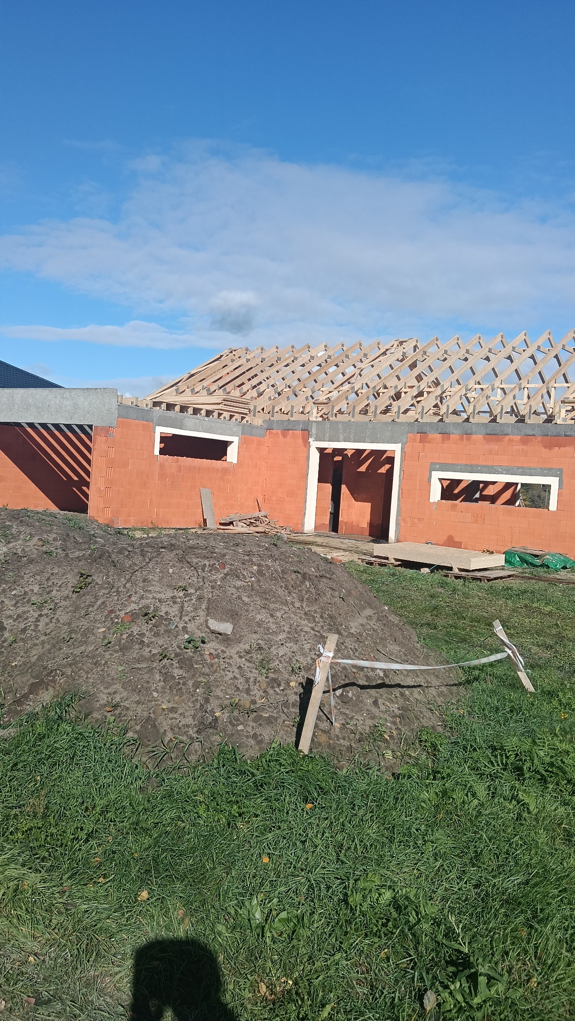 Surowy stan domu z czerwonej cegły z drewnianą konstrukcją dachu w trakcie budowy, ziemia na pierwszym planie, błękitne niebo w tle.