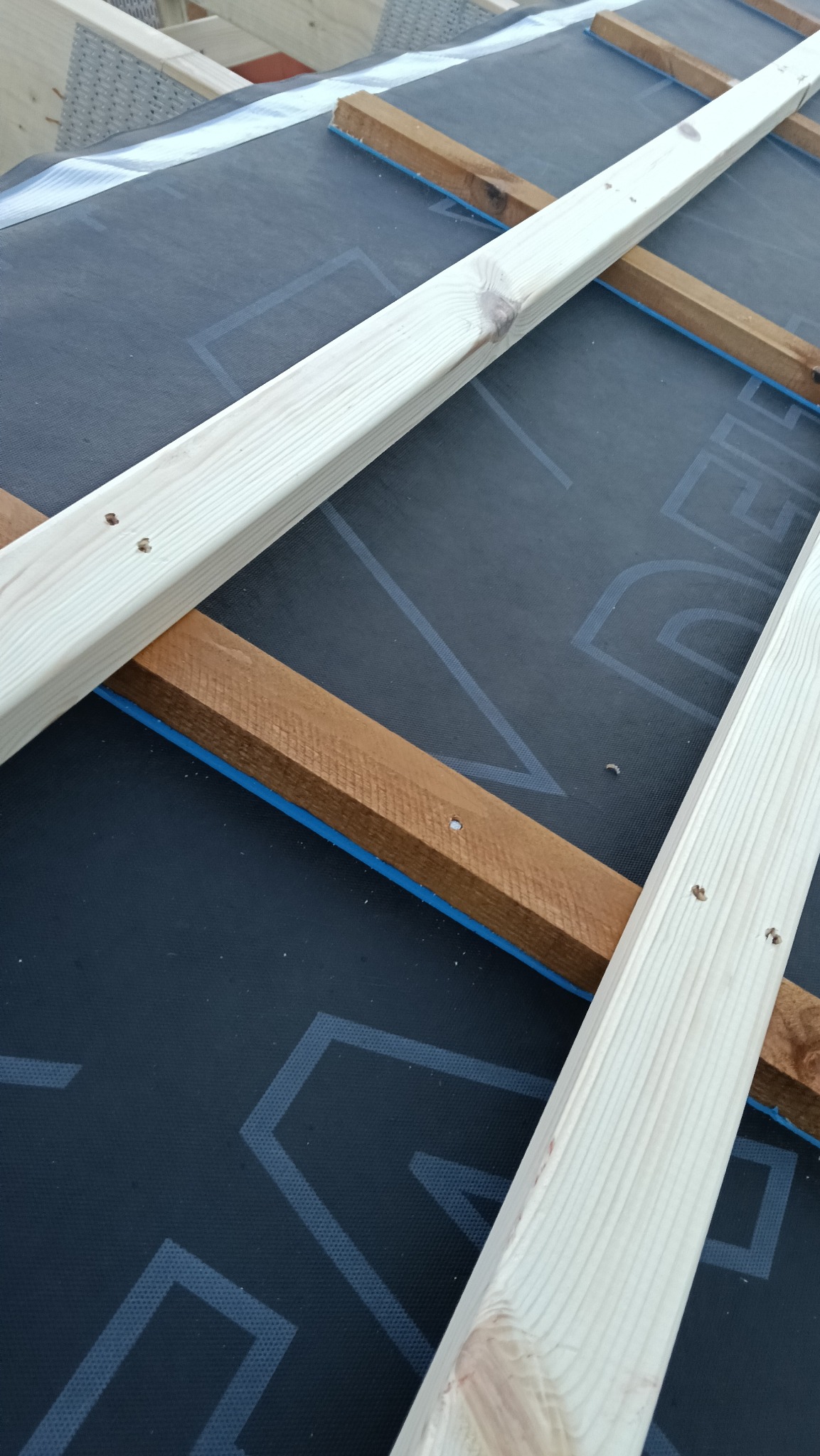 Konstrukcja dachu w trakcie budowy: czarna membrana z nadrukiem, drewniane łaty w kolorze jasnego drewna i brązu, mocowane do membrany z użyciem niebieskiej taśmy.