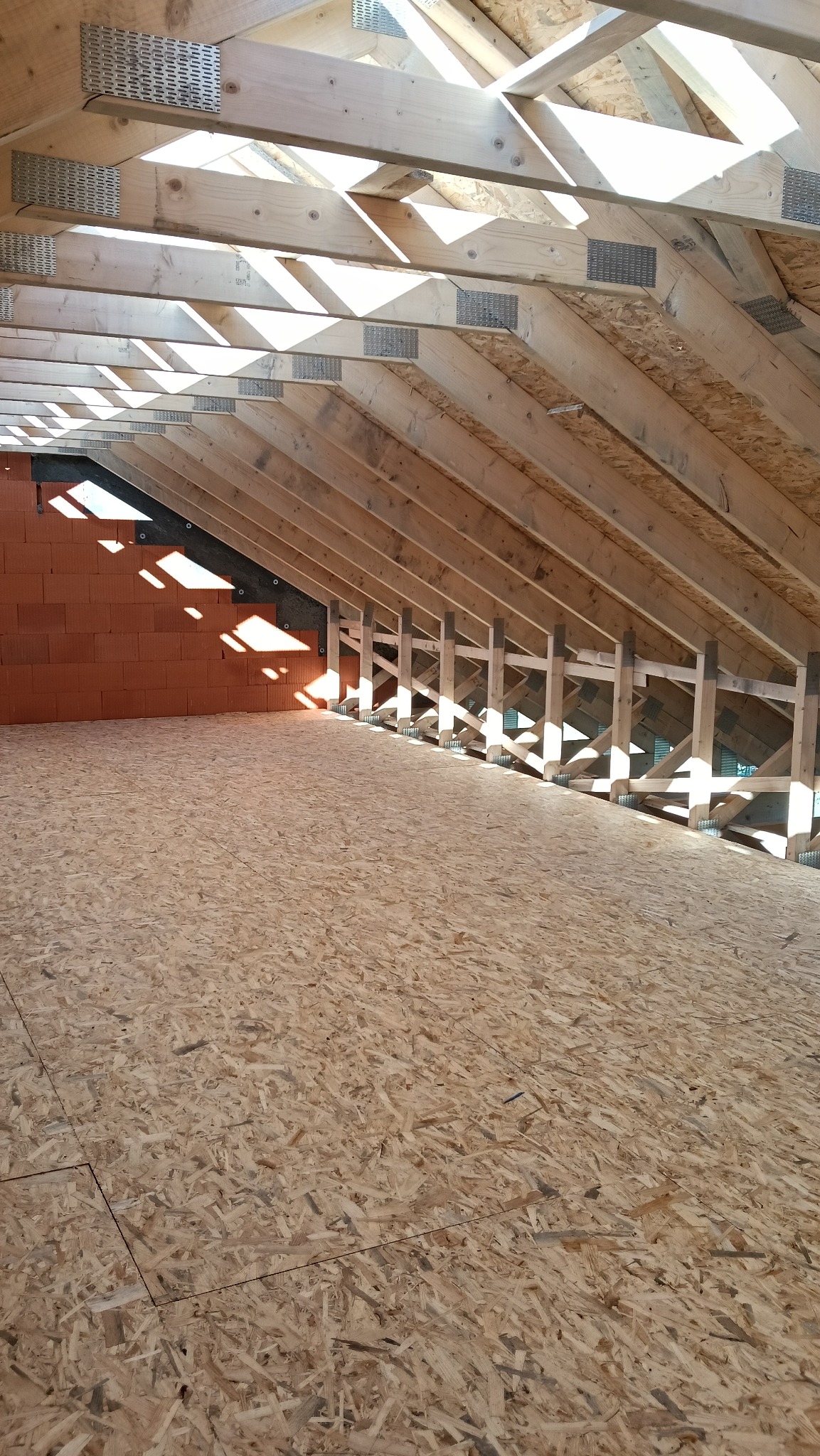 Konstrukcja drewniana dachu z widocznymi elementami złącznymi i fragmentem ściany z pustaków ceramicznych. Podłoga wyłożona płytami OSB. Widoczne promienie słoneczne.