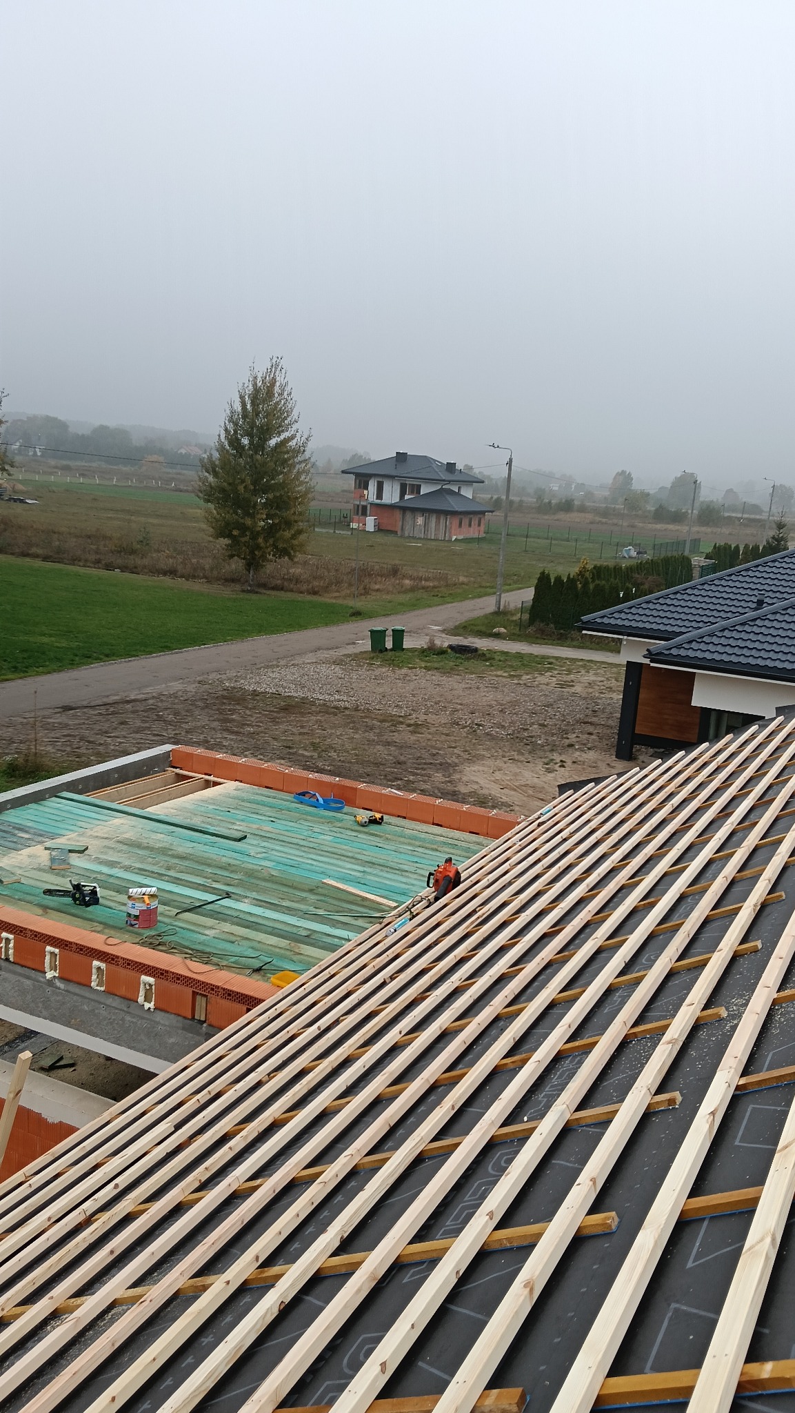 Wysoki kąt: konstrukcja dachu z drewnianymi łatami na czarnej membranie, narzędzia na zielonej płycie OSB. W tle domy i drzewa we mgle.