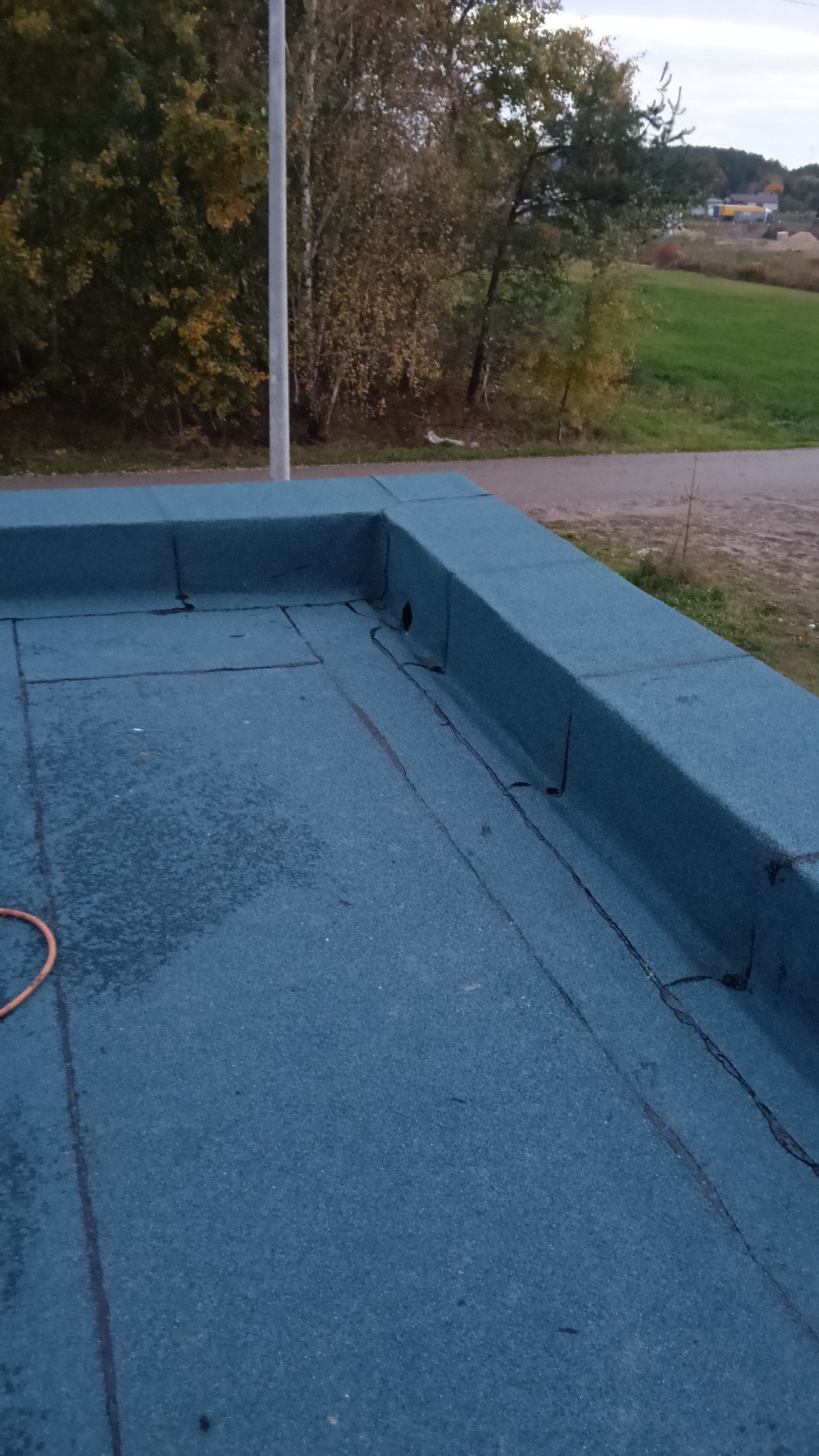 Niebieski, płaski dach pokryty papą z widocznym odpływem wody, otoczony obróbką blacharską. W tle drzewa i słup oświetleniowy.