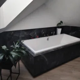 Nowoczesna łazienka na poddaszu z wanną obudowaną czarnym marmurem, drewnopodobna podłoga, roślina doniczkowa. Stonowana kolorystyka, minimalistyczny design.