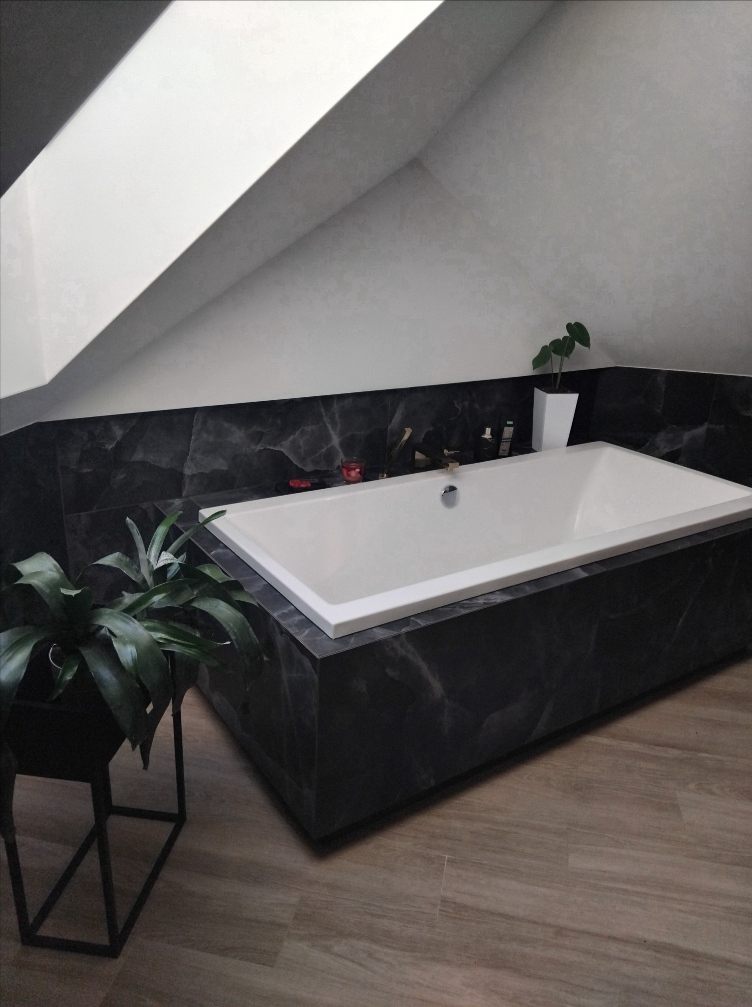 Nowoczesna łazienka na poddaszu z wanną obudowaną czarnym marmurem, drewnopodobna podłoga, roślina doniczkowa. Stonowana kolorystyka, minimalistyczny design.
