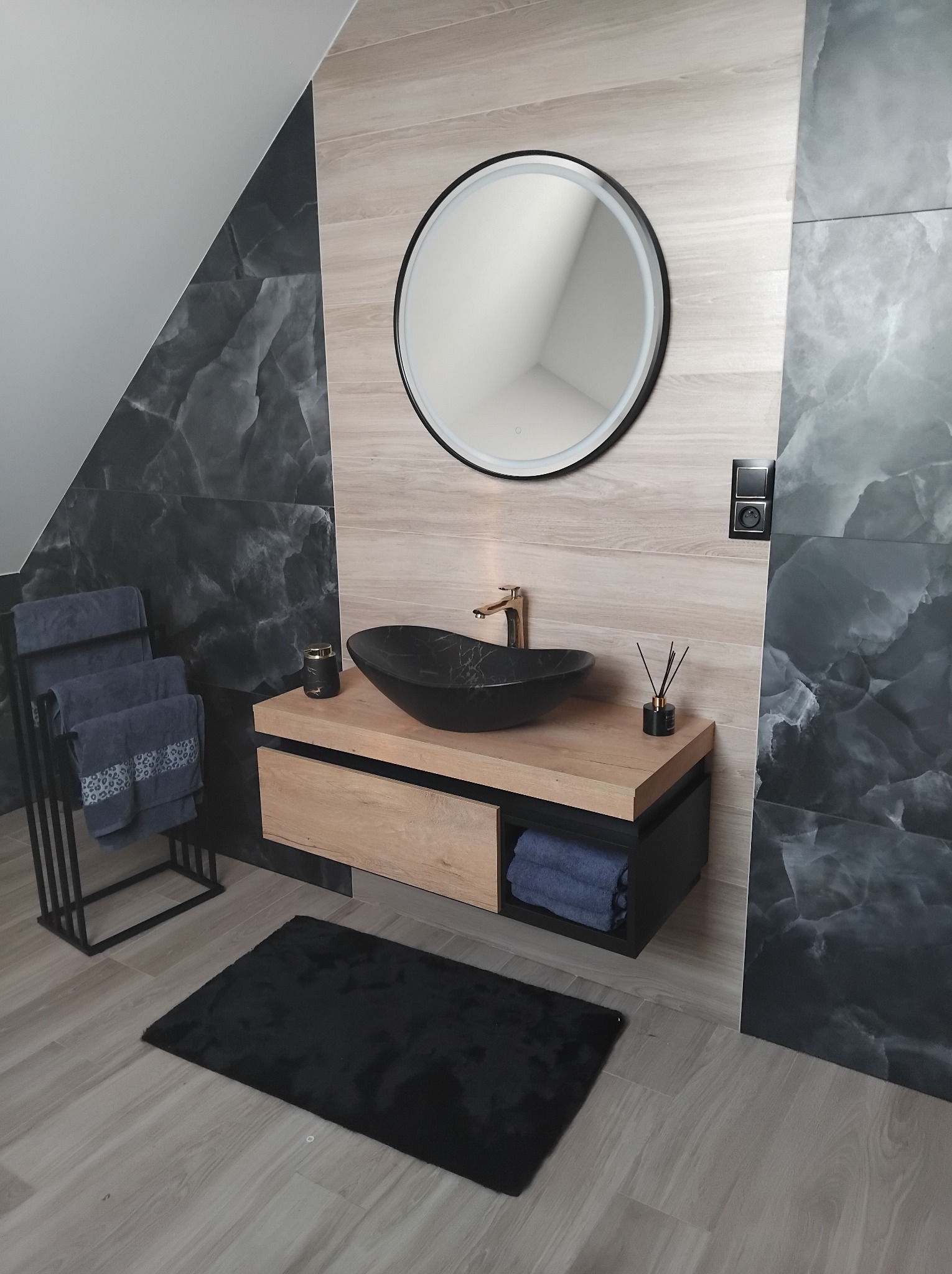 Nowoczesna łazienka na poddaszu: czarna umywalka na drewnianej szafce, okrągłe lustro z podświetleniem, szare płytki imitujące marmur i czarny dywanik.