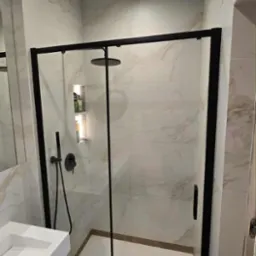 Nowoczesna kabina prysznicowa z czarną ramą i deszczownicą, wykończona marmurowymi płytkami. Umywalka z białego konglomeratu.