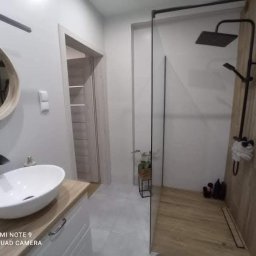 Usługi Remontowo-Wykonczeniowe Kamil Klunder - Nowoczesna łazienka z kabiną prysznicową typu walk-in, okrągłym lustrem i umywalką nablatową. Drewnopodobne płytki na ścianie i podłodze.