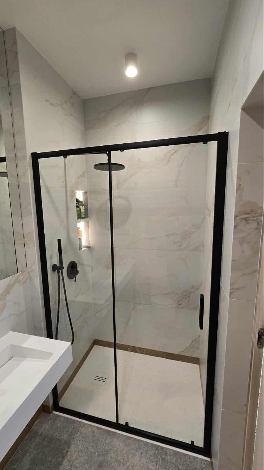 Nowoczesna kabina prysznicowa z czarną ramą i deszczownicą, wykończona marmurowymi płytkami. Umywalka z białego konglomeratu.