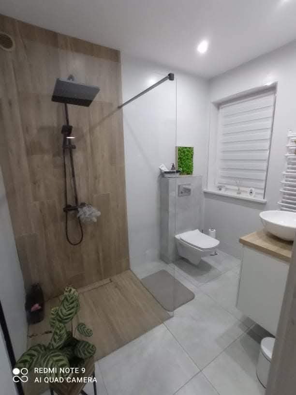 Nowoczesna łazienka po remoncie: kabina typu walk-in z deszczownicą, drewnopodobne płytki, biała ceramika, rośliny doniczkowe. Minimalistyczny design.