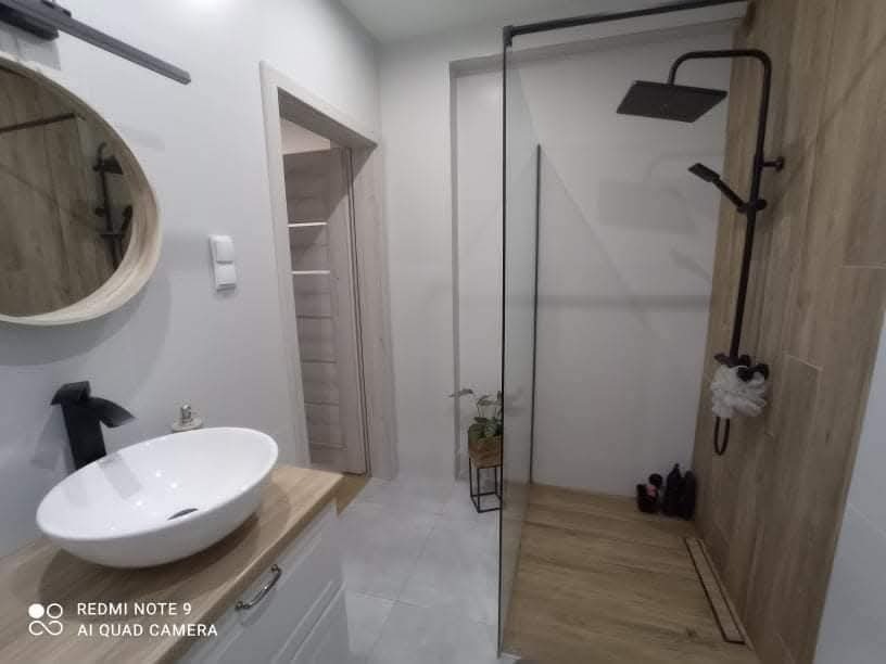 Nowoczesna łazienka z kabiną prysznicową typu walk-in, okrągłym lustrem i umywalką nablatową. Drewnopodobne płytki na ścianie i podłodze.