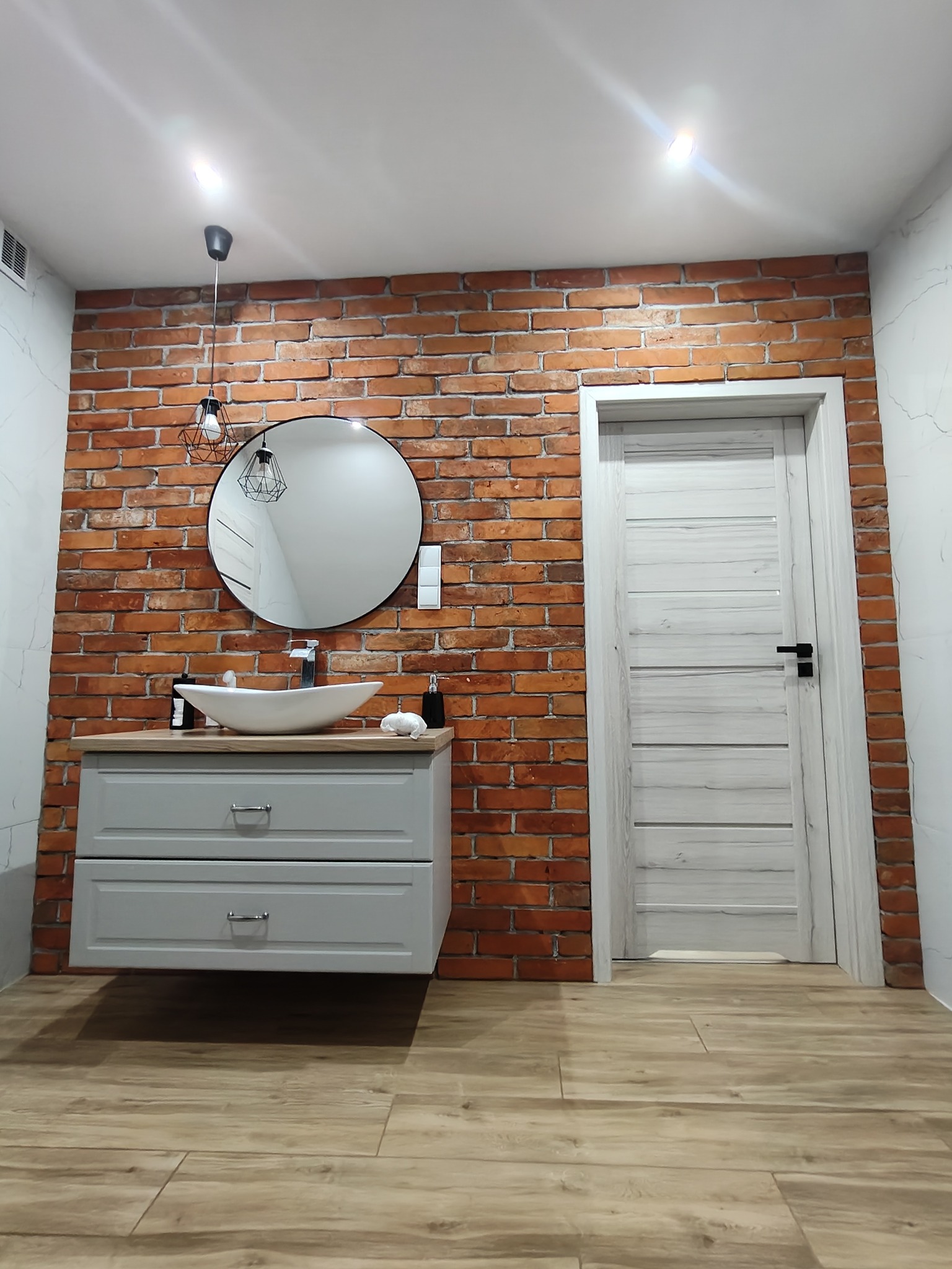 Nowoczesna łazienka w Czersku z umywalką na szafce, okrągłym lustrem i ścianą z cegły. Podłoga wyłożona panelami imitującymi drewno. Stylowe oświetlenie.