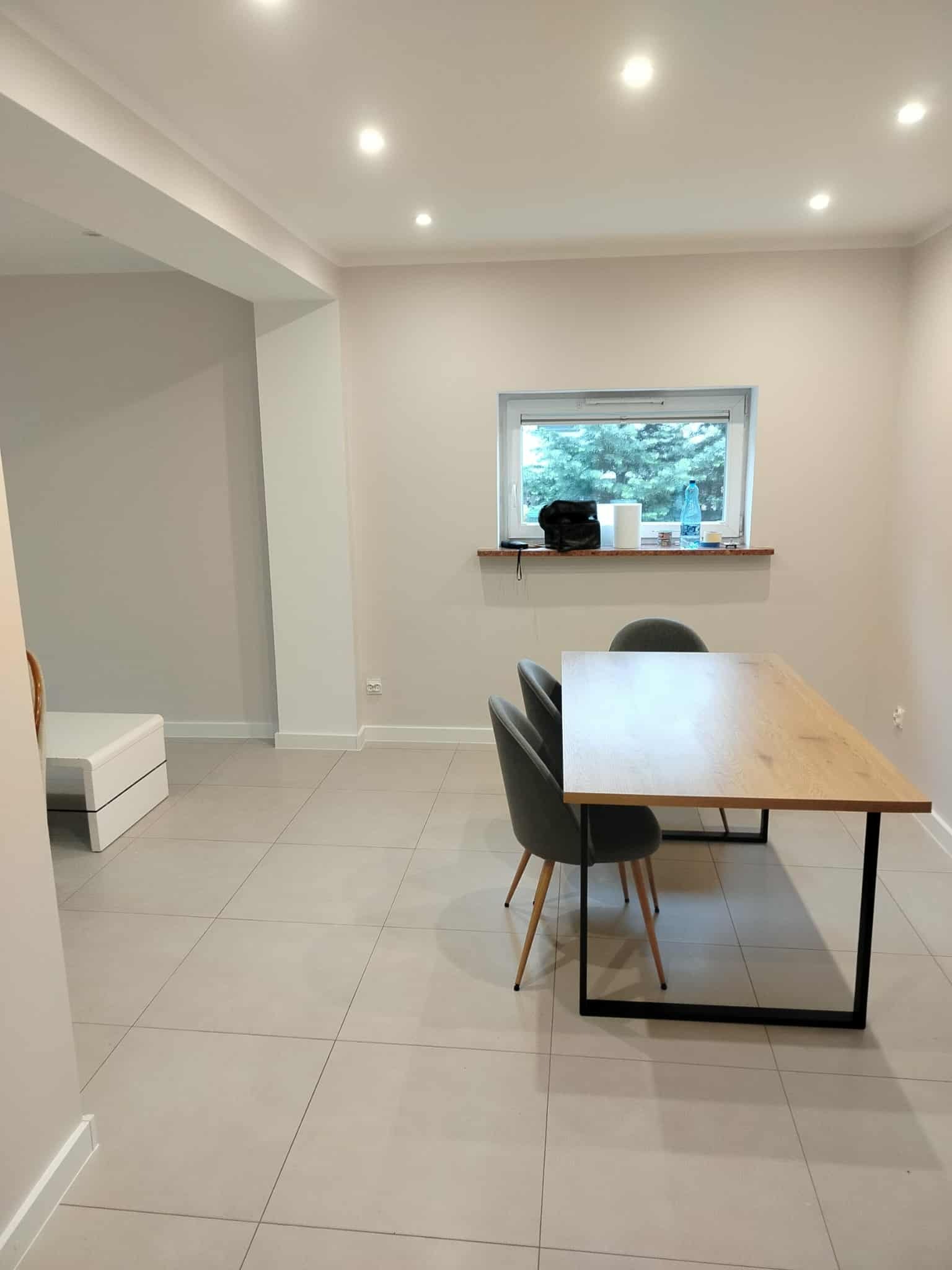 Jasne wnętrze po remoncie: stół z krzesłami na tle okna. Widoczne świeżo położone płytki podłogowe i pomalowane ściany. Minimalistyczny design.