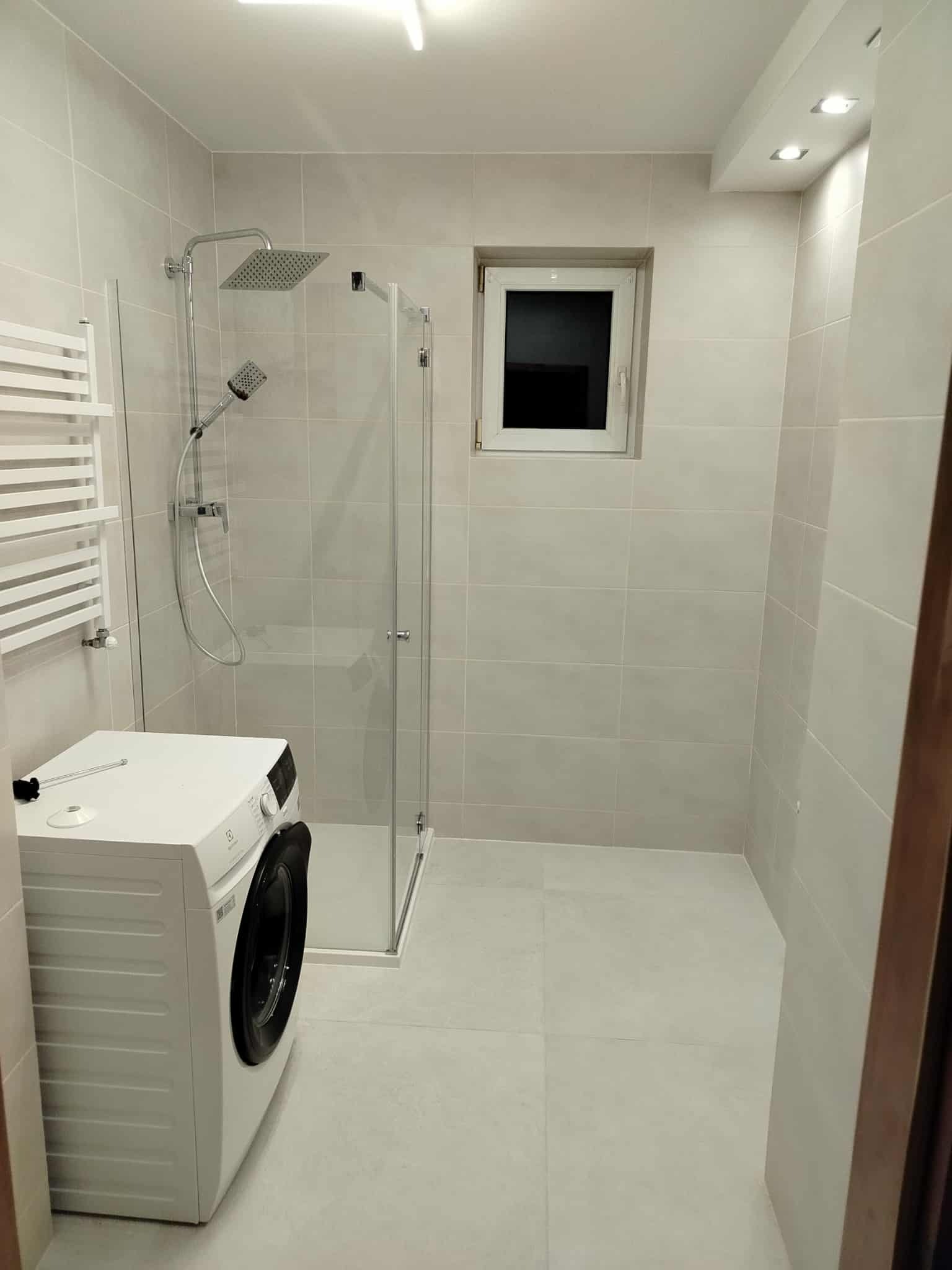 Jasna, wykafelkowana łazienka z kabiną prysznicową, grzejnikiem drabinkowym i pralką. Małe okno z białymi ramami. Minimalistyczny design.