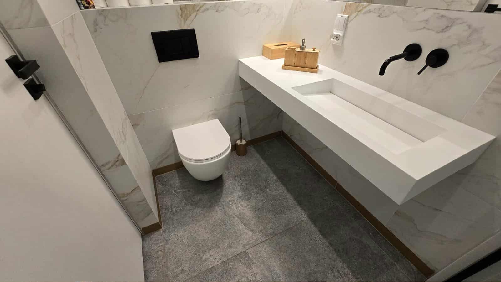 Nowoczesna łazienka po remoncie: biała ceramika, marmurowe płytki i szara podłoga. Umywalka z czarną armaturą, toaleta podwieszana.