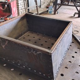Stal Tech Construct - Metalowa skrzynia z perforowanym dnem, spawana punktowo na stole spawalniczym. Widoczne narzędzia i ślady obróbki metalu.