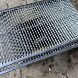 Dwie metalowe kraty z równoległymi prętami, ułożone jedna na drugiej na szarym brukowanym podłożu. Widoczne spawy i precyzyjne wykonanie.