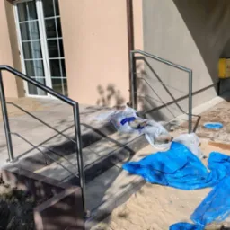 Nierdzewna, nowoczesna poręcz schodowa z metalowymi słupkami, zamontowana przy wejściu do domu. Widoczne materiały budowlane i elementy wykończeniowe.