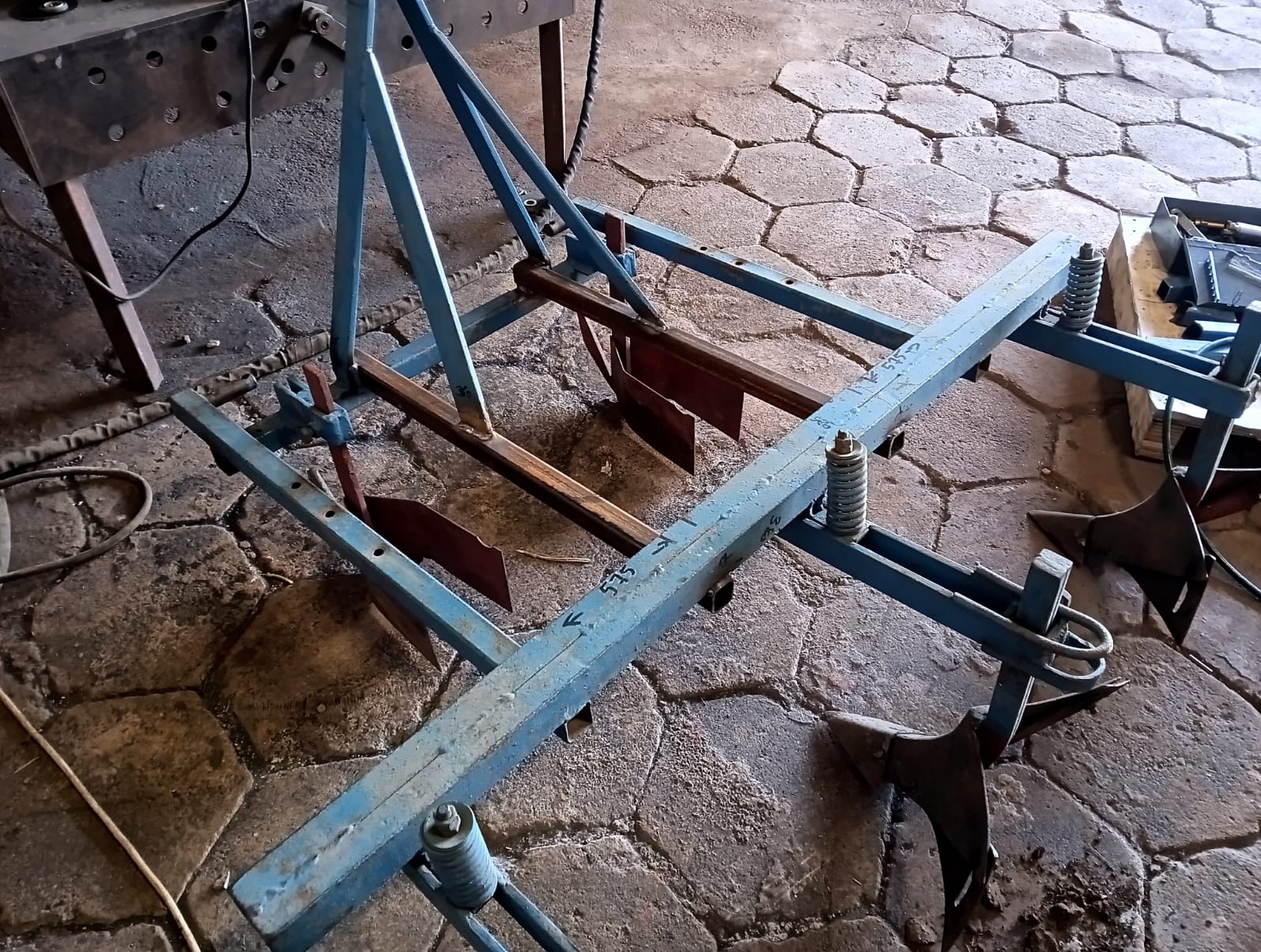 Niebieska, spawana konstrukcja metalowa z elementami roboczymi, leżąca na posadzce z kamiennej kostki. Widoczne ślady spawania i oznaczenia na metalu.