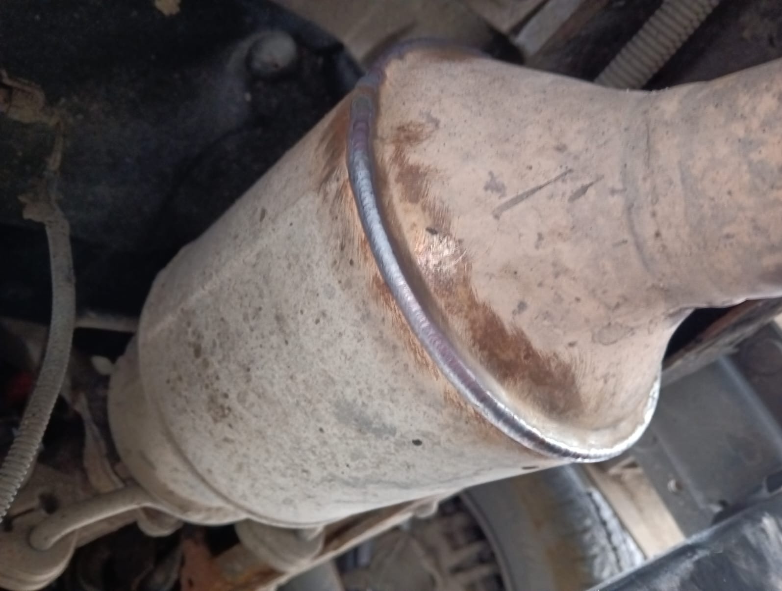 Spawany tłumik samochodowy z widocznym świeżym spawem, lekko zabrudzony, zamontowany pod podwoziem pojazdu. Widok z dołu, perspektywa ukośna.