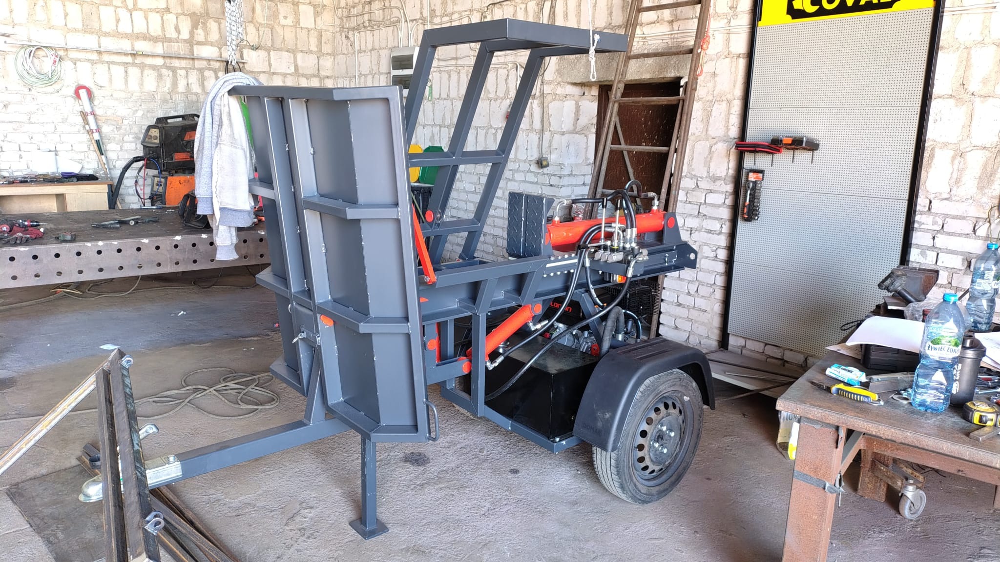 Szara, metalowa konstrukcja z elementami hydrauliki na przyczepce, spawana w warsztacie. W tle ceglana ściana i narzędzia.