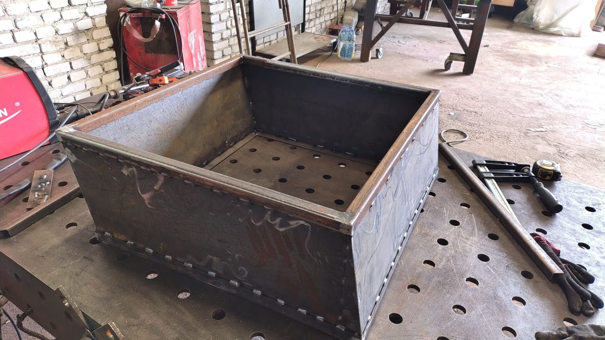 Metalowa skrzynia z perforowanym dnem, spawana punktowo na stole spawalniczym. Widoczne narzędzia i ślady obróbki metalu.