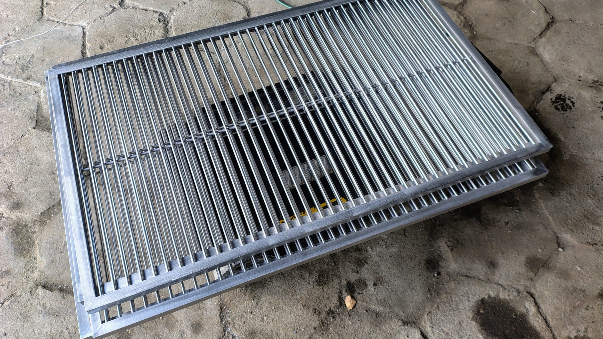 Dwie metalowe kraty z równoległymi prętami, ułożone jedna na drugiej na szarym brukowanym podłożu. Widoczne spawy i precyzyjne wykonanie.