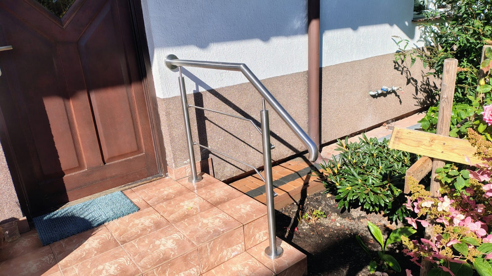 Nierdzewna, prosta poręcz przy schodach wejściowych do domu z brązowymi drzwiami. Widoczne płytki na schodach i fragment ogrodu z kwiatami.