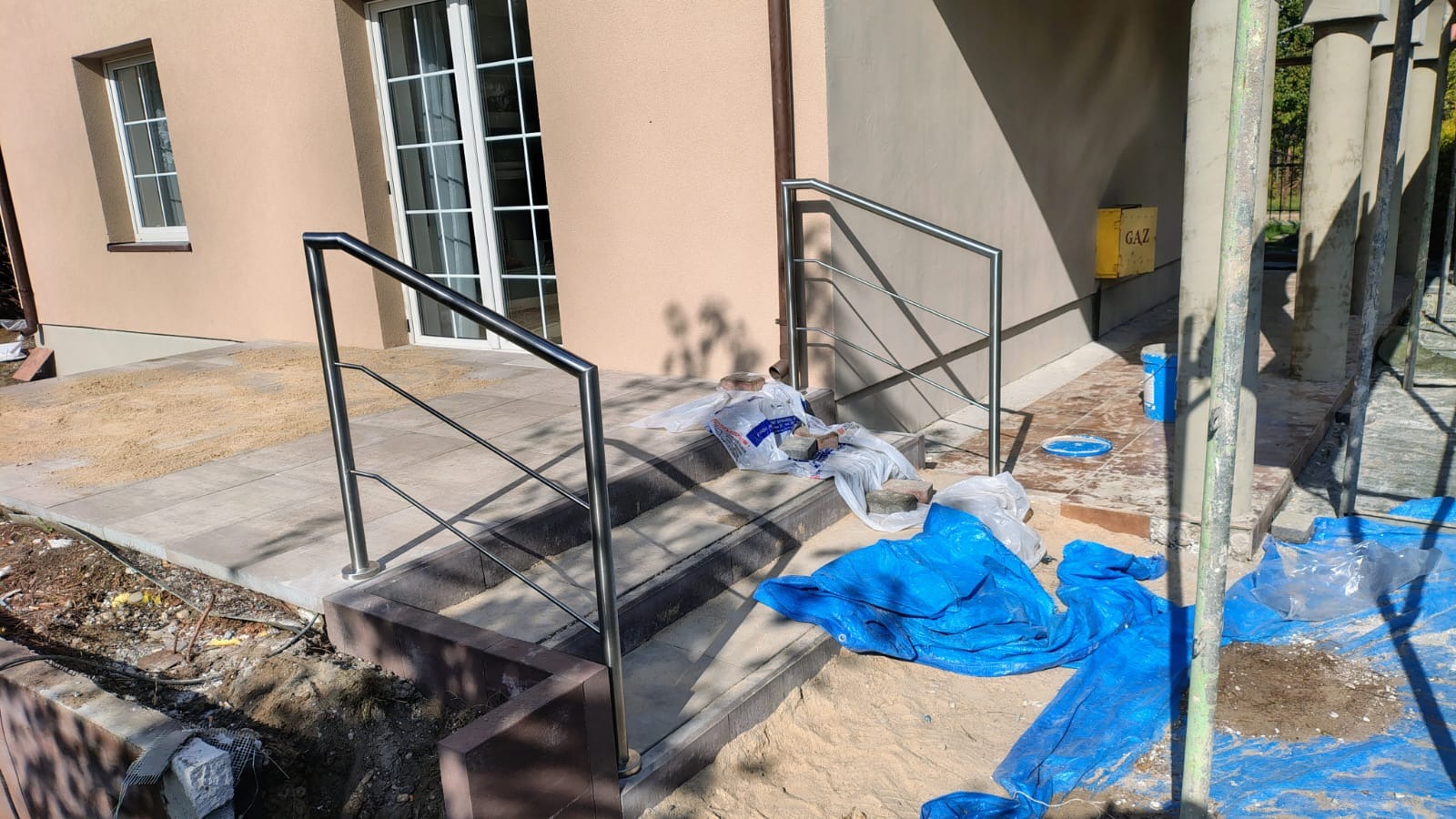 Nierdzewna, nowoczesna poręcz schodowa z metalowymi słupkami, zamontowana przy wejściu do domu. Widoczne materiały budowlane i elementy wykończeniowe.