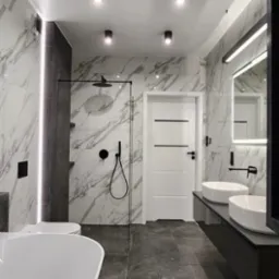 Elegancka łazienka z marmurowymi płytkami, czarną armaturą i podwieszanymi umywalkami. Oświetlenie punktowe podkreśla nowoczesny design i wysoką jakość wykończenia.