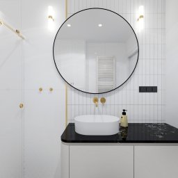 Corners Pracownia Paweł Szustakowski - Elegancka łazienka z okrągłym lustrem i czarnym blatem umywalki. Białe płytki na ścianach i podłodze z geometrycznym wzorem. Złote akcenty armatury i oświetlenia.