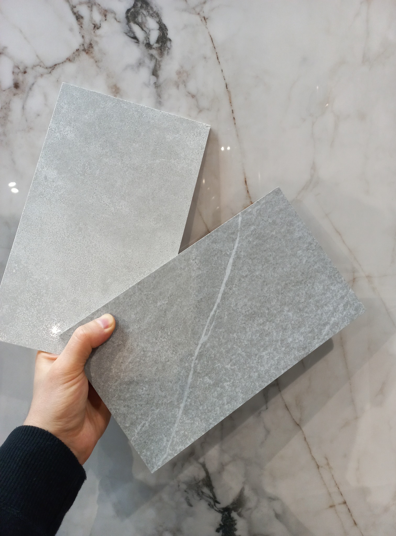 Dłoń prezentuje dwie płytki ceramiczne w odcieniach szarości na tle marmurowej ściany. Jedna płytka gładka, druga z subtelnym wzorem żyłek.