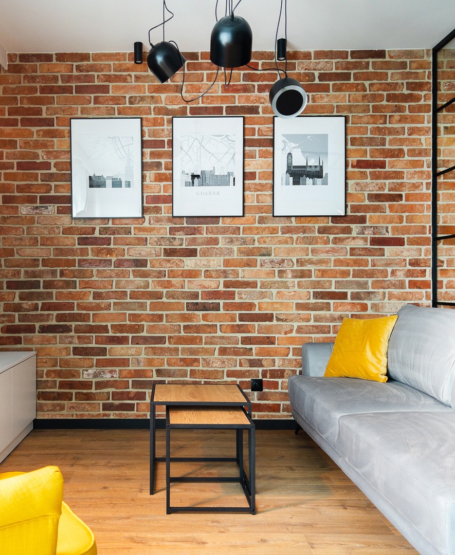 Ceglana ściana z trzema grafikami w czarnych ramkach, czarne lampy wiszące, szara sofa z żółtą poduszką i drewniana podłoga. Widok na salon w stylu industrialnym.