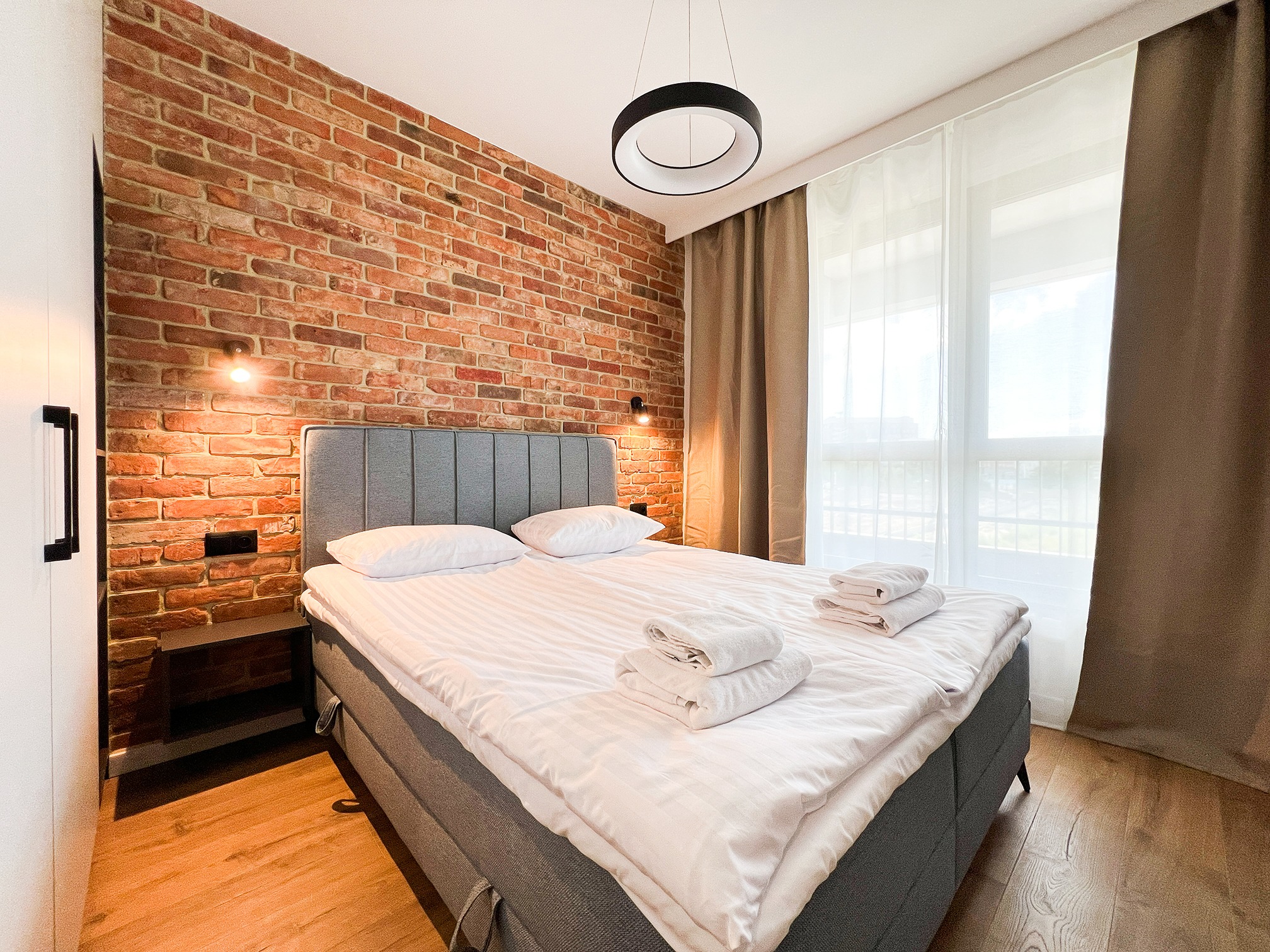 Sypialnia z ceglaną ścianą, szarym łóżkiem i okrągłą lampą. Widoczne drewniane panele podłogowe i zasłony w oknie. Nowoczesny design z elementami rustykalnymi.