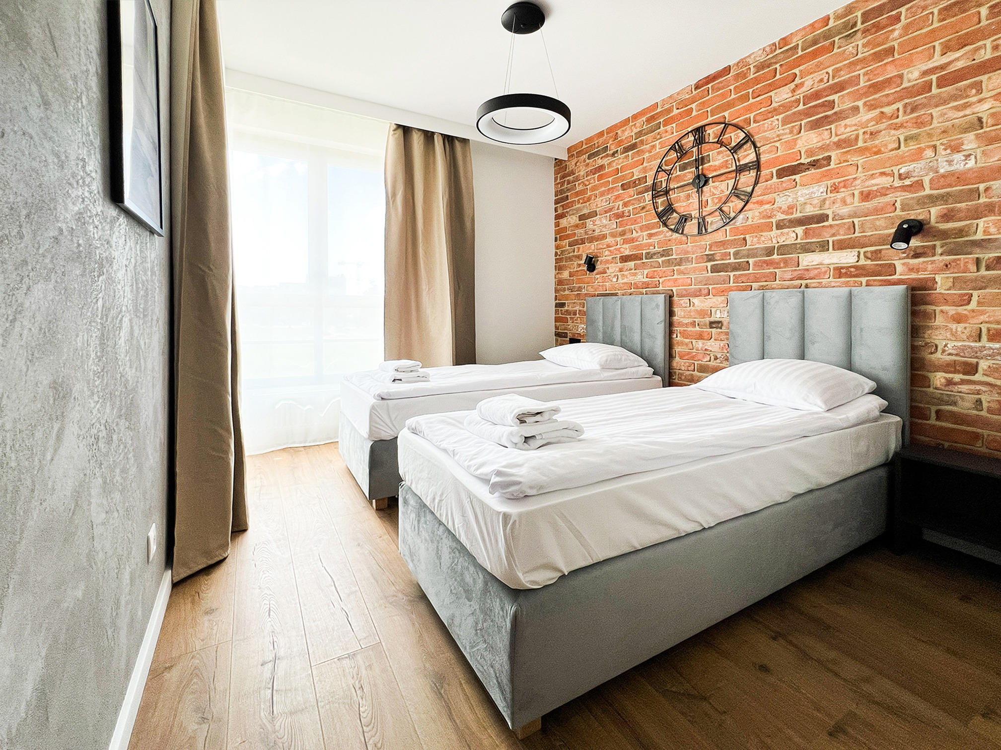 Pokój z dwoma łóżkami pojedynczymi, ceglaną ścianą z zegarem i nowoczesnym oświetleniem. Widok na okno z zasłonami i drewnianą podłogę.