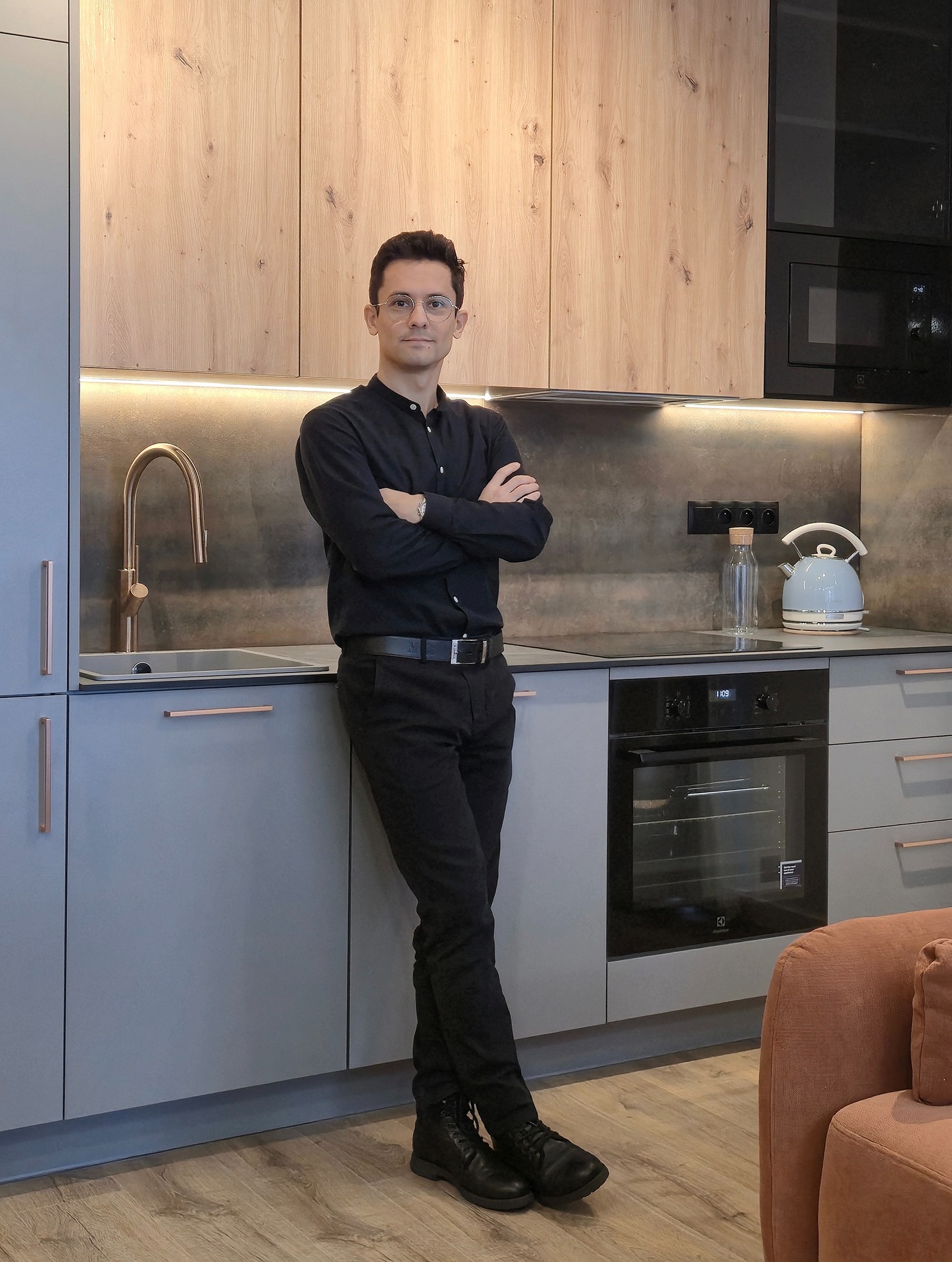 Mężczyzna w czerni na tle nowoczesnej kuchni z szarymi szafkami, drewnianymi frontami górnymi i złotą armaturą. Widoczny piekarnik i płyta indukcyjna.