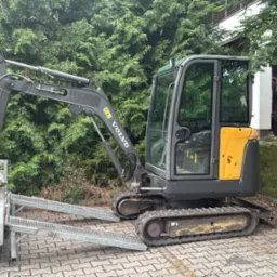 Szara minikoparka Volvo na przyczepie z najazdami, z łyżką załadowaną ziemią, na tle zieleni i budynku. Sprzęt gotowy do transportu.