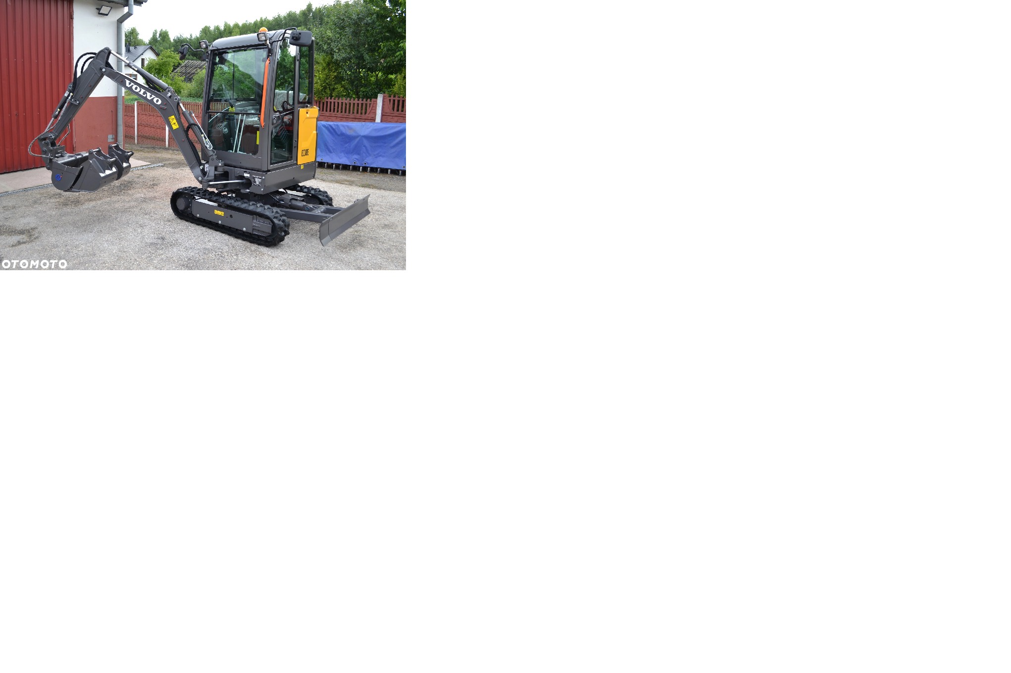 Szara minikoparka Volvo z łyżką i lemieszem na żwirowym podłożu, z widocznym logo Volvo na ramieniu. Tło stanowi budynek i zieleń.